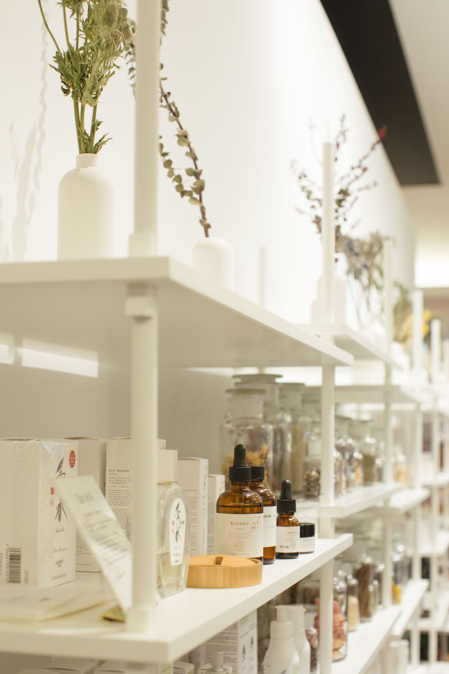White shelves display glass jars, bottles and dried flowers in a minimalist apothecary or wellness store interior.
