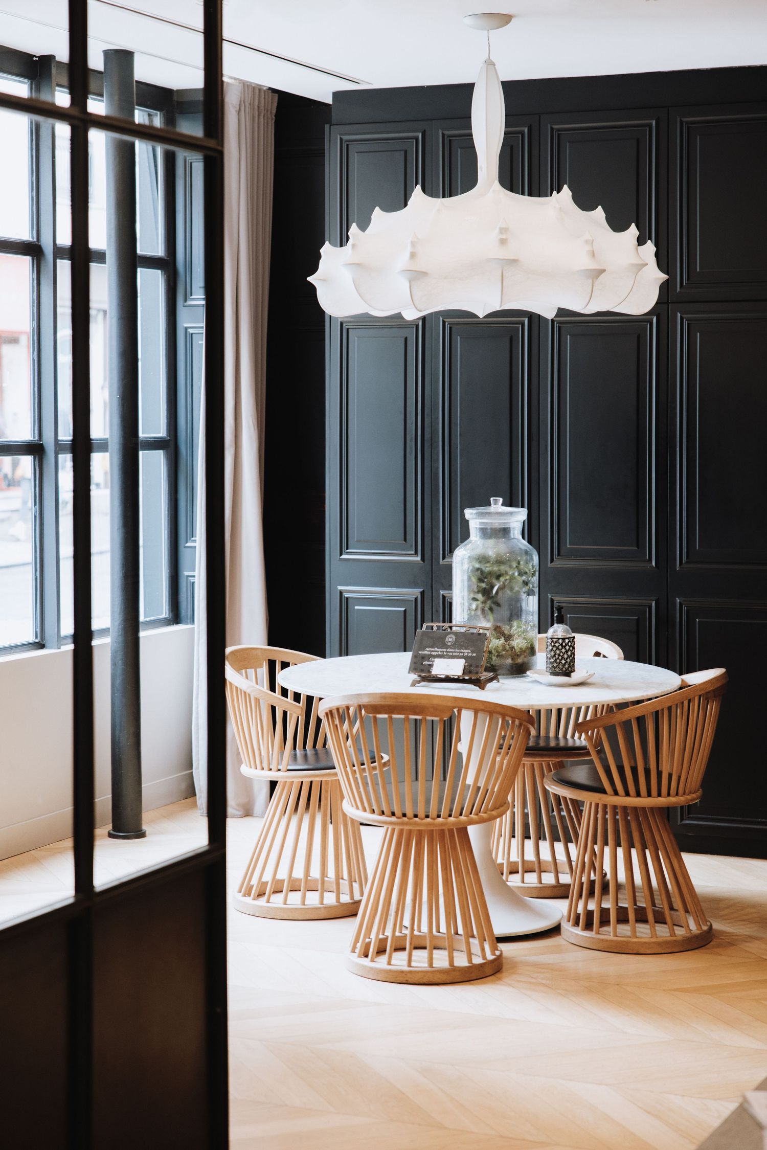 Modern dining area with round white table, wooden spindle chairs, and dramatic white pendant light against dark paneled walls.