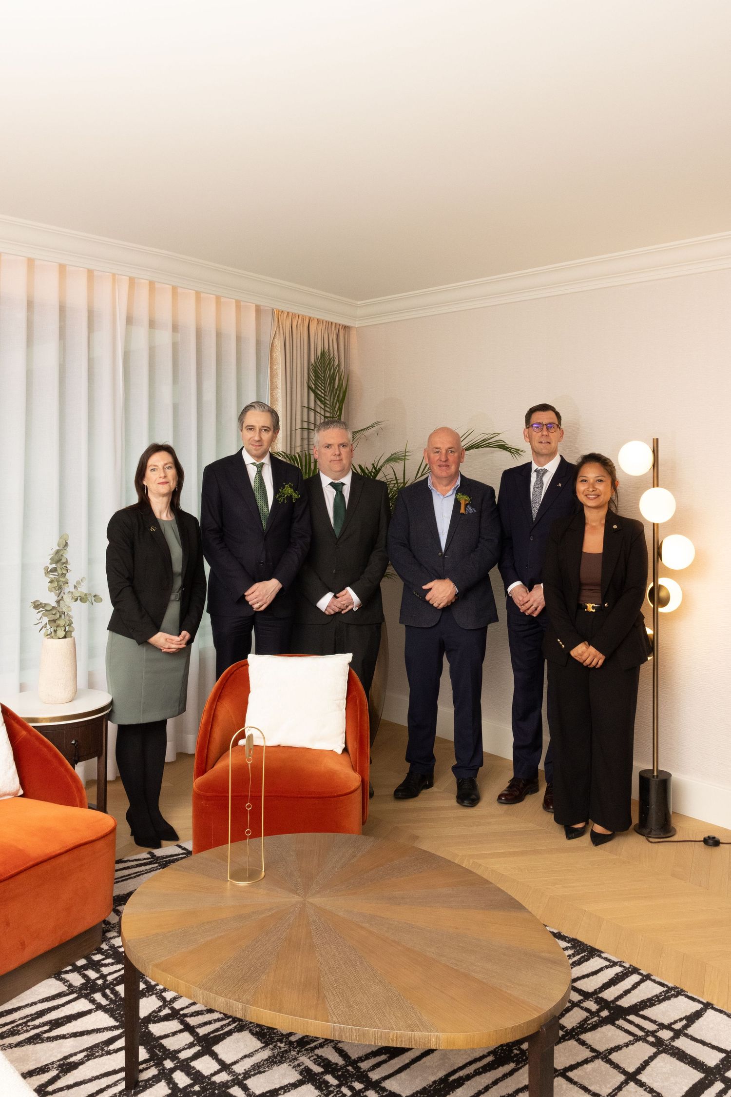 A group of formally dressed people standing in a modern room with a wooden table and patterned rug.