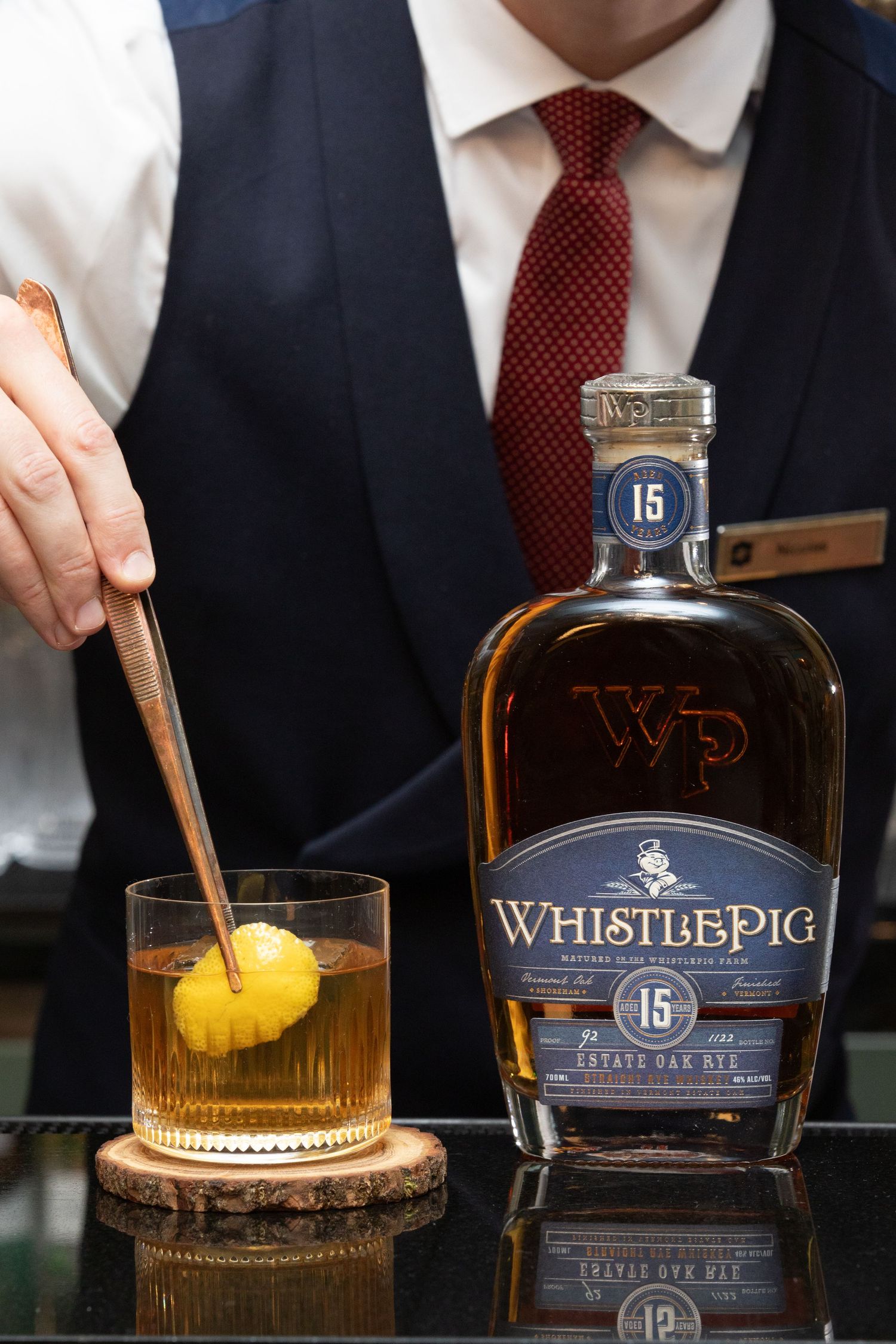 A bottle of WhistlePig aged whiskey sits beside a glass with an orange garnish and ice cube on a dark surface.