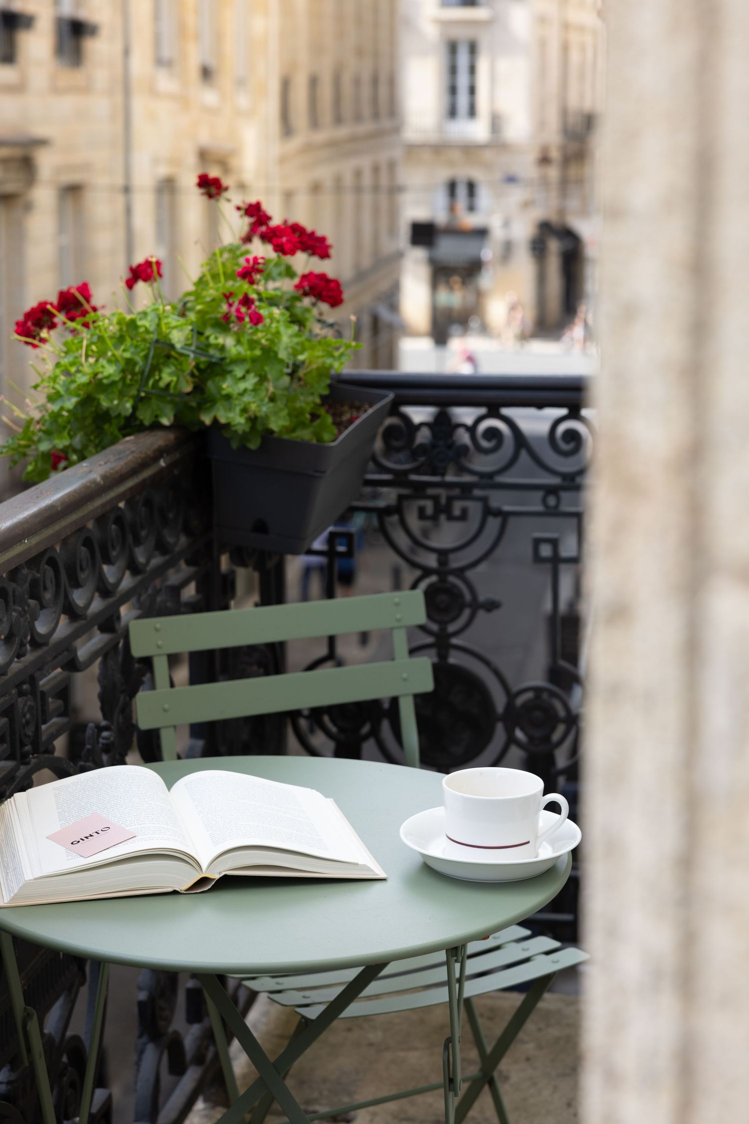 A cozy Parisian balcony with red geraniums, an open book, and teacup on a mint green bistro table and chair.