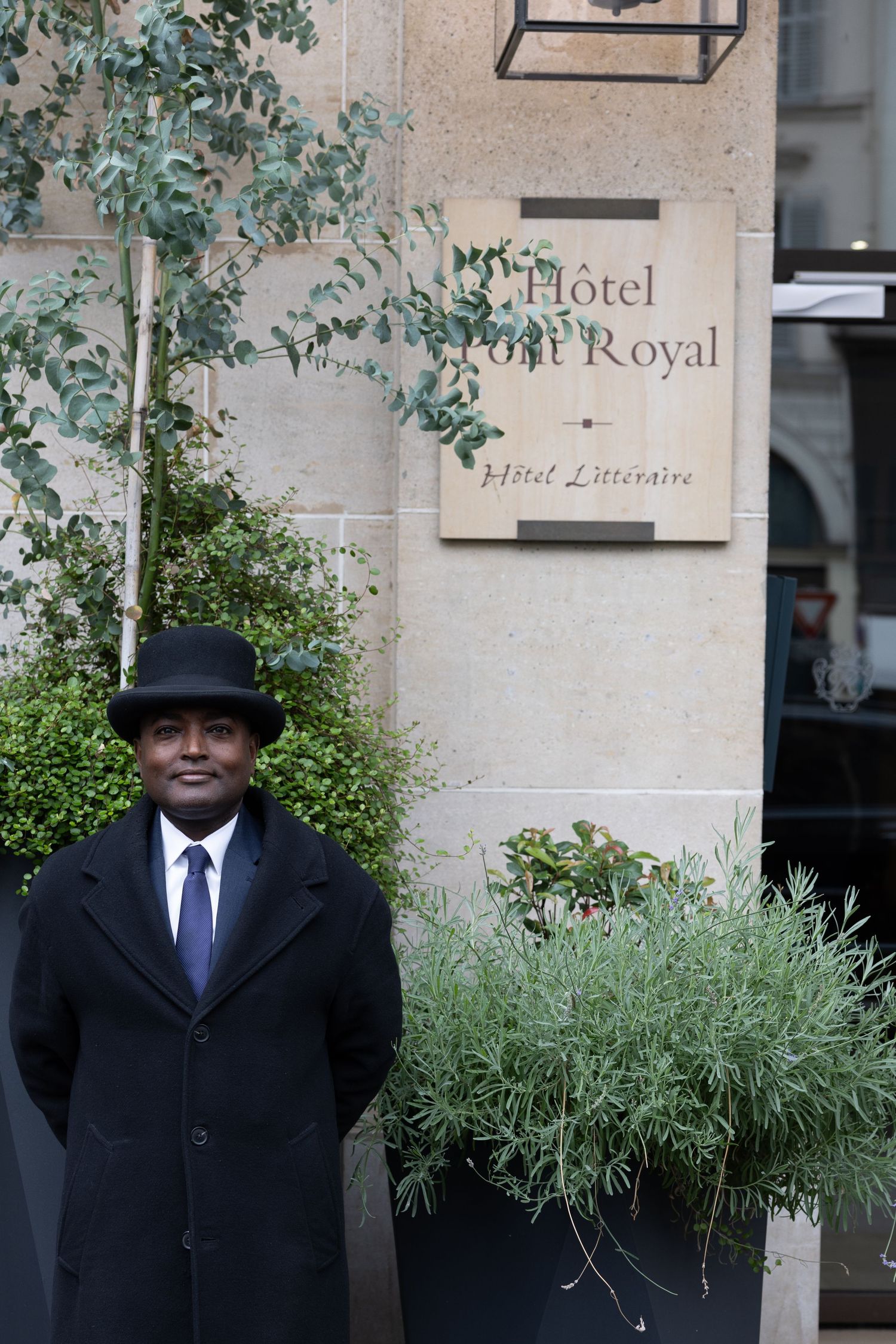 A figure in a black coat and hat stands outside the entrance of a hotel with green plants in the foreground.