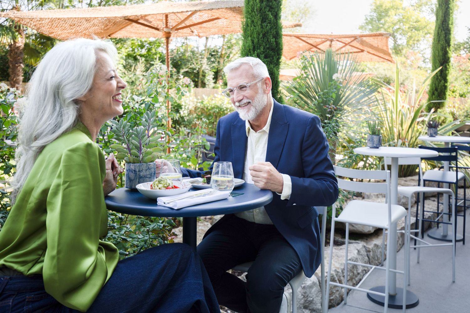 Two people enjoying coffee and conversation at an outdoor cafe patio with orange umbrellas and greenery.
