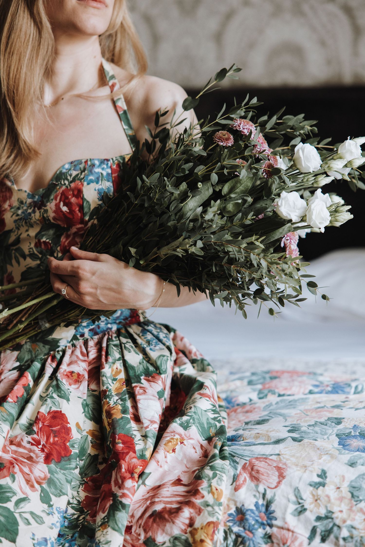 Person in floral dress arranges fresh greenery and flowers on white surface with artistic styling.