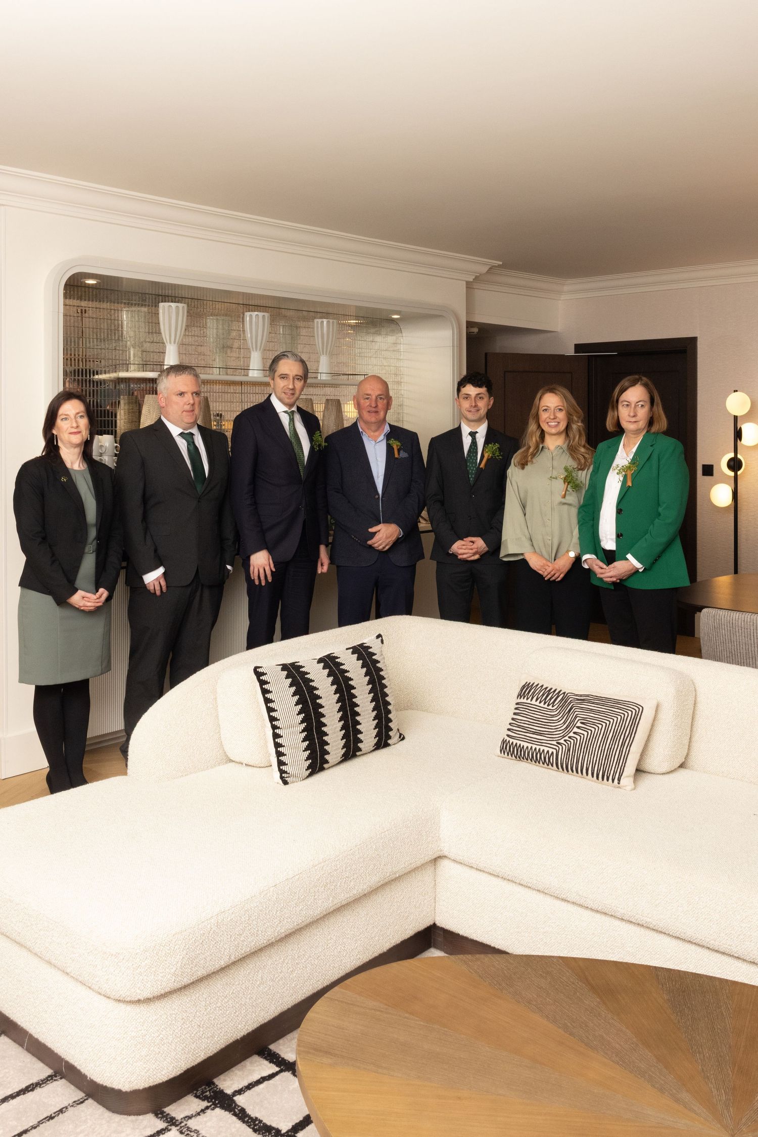 A group of professionally dressed people stand in a modern living room with a white sectional sofa and decorative pillows.