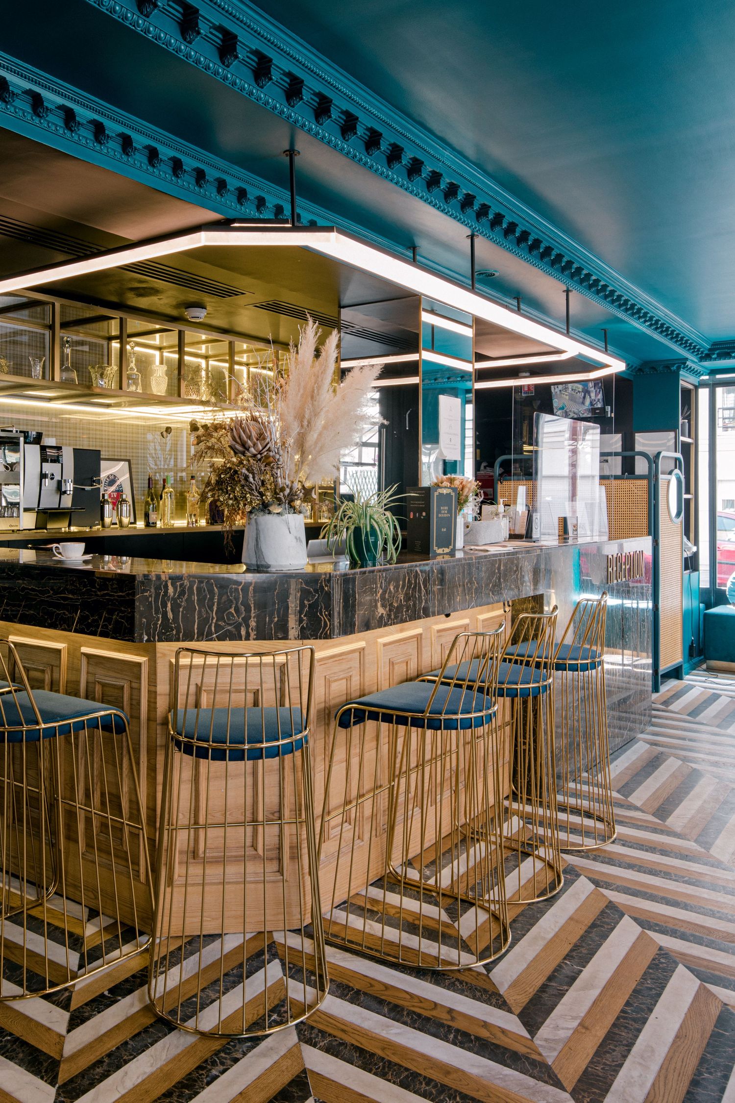 Modern restaurant bar interior with chevron patterned floor, gold barstools and teal accents under decorative lighting.