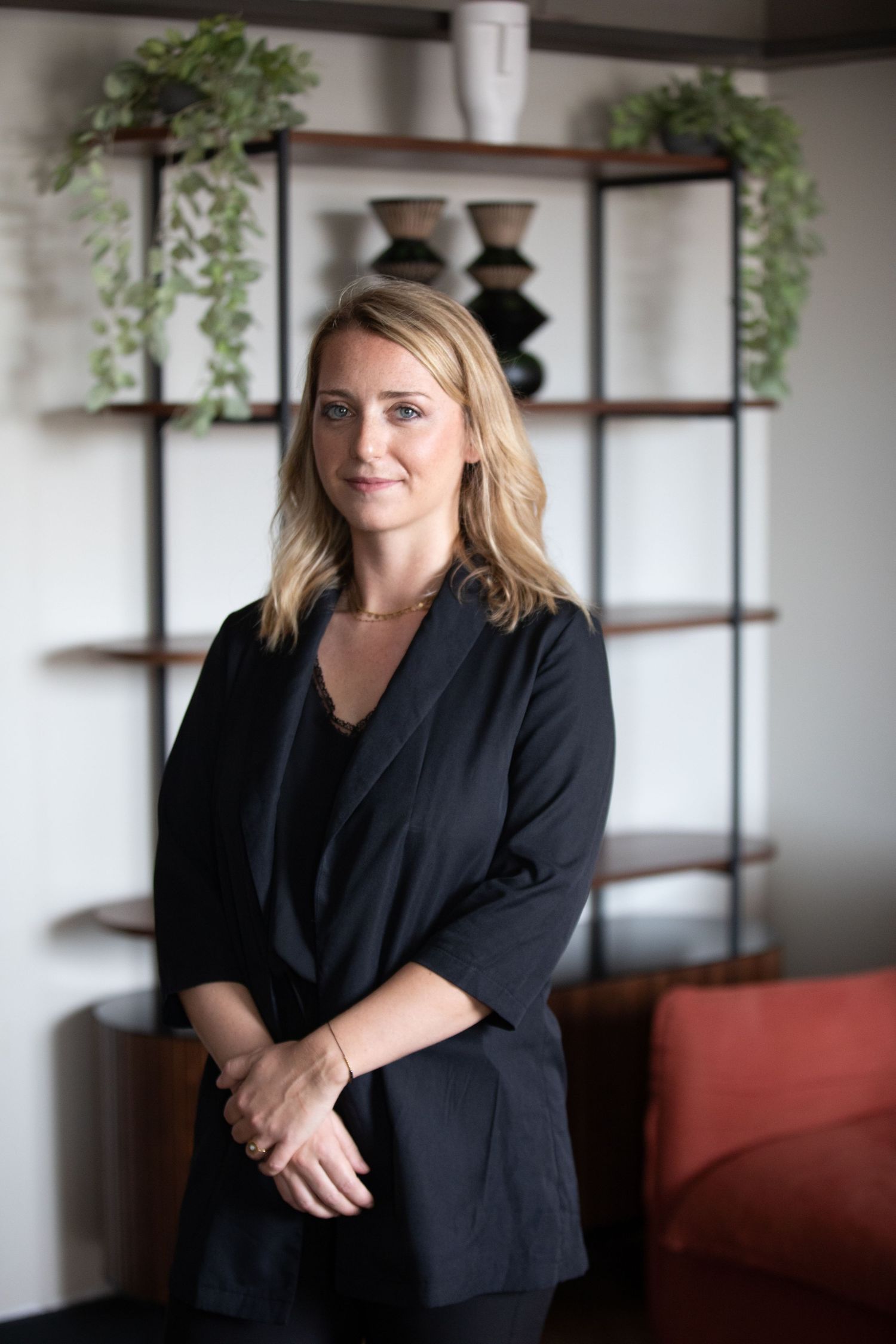 Professional portrait in an office setting with wooden shelving, plants, and modern decor in the background.