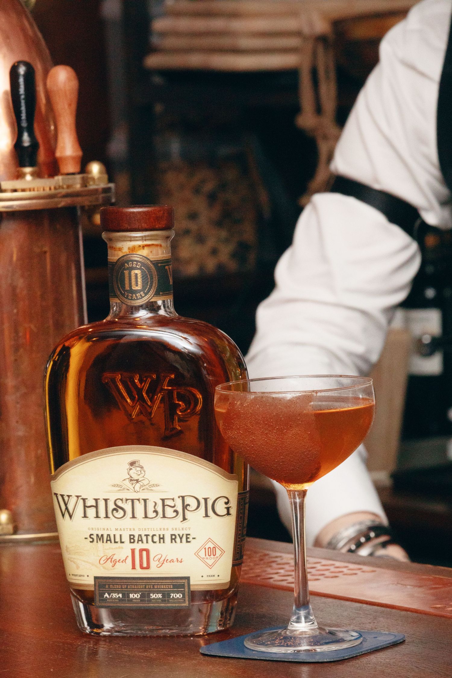 A glass of whiskey sits on a bar counter next to bottles of WhistlePig rye whiskey in a dimly lit setting.