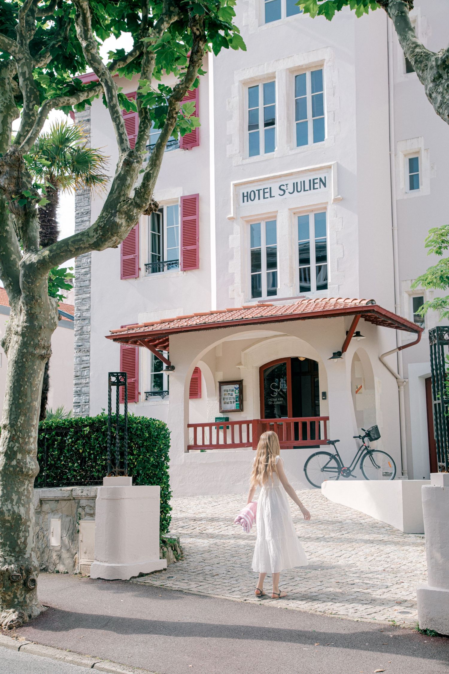 A charming white Hotel St-Julien building with red shutters and a covered entrance porch in a quaint European setting.