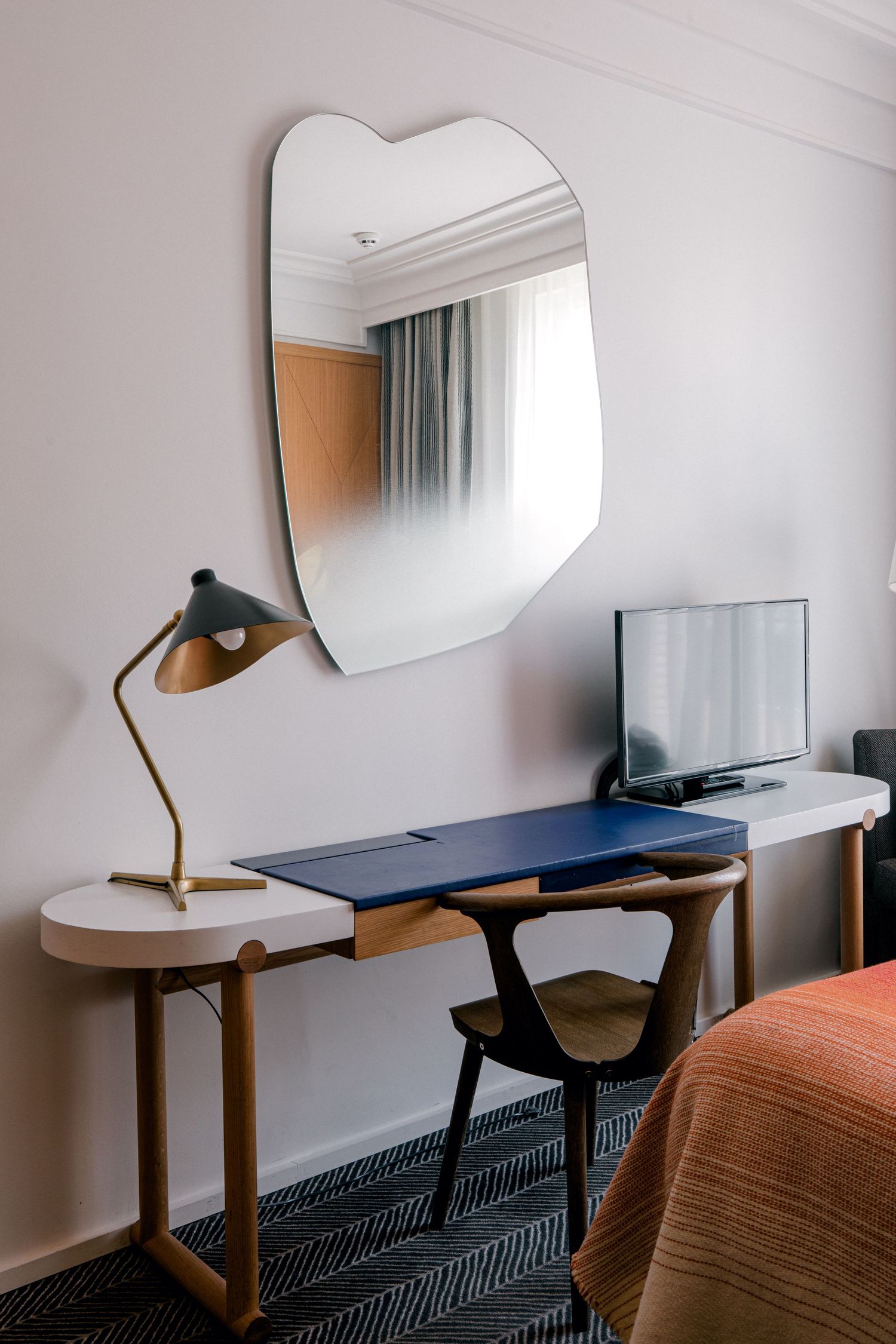 Modern hotel room workspace with curved mirror, brass desk lamp, navy blue desk surface and flat screen TV on white wall.