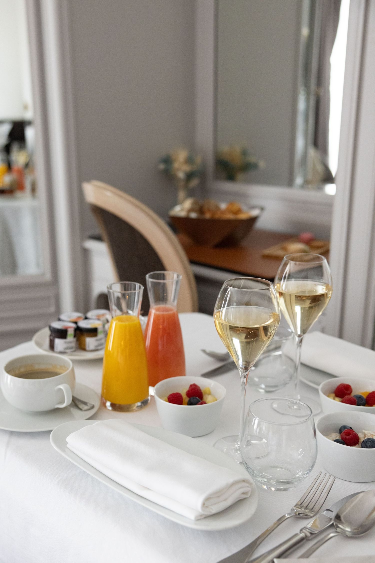 Elegant breakfast table setting with wine glasses, fresh juice carafes, coffee cup, and bowls of berries on white tablecloth.