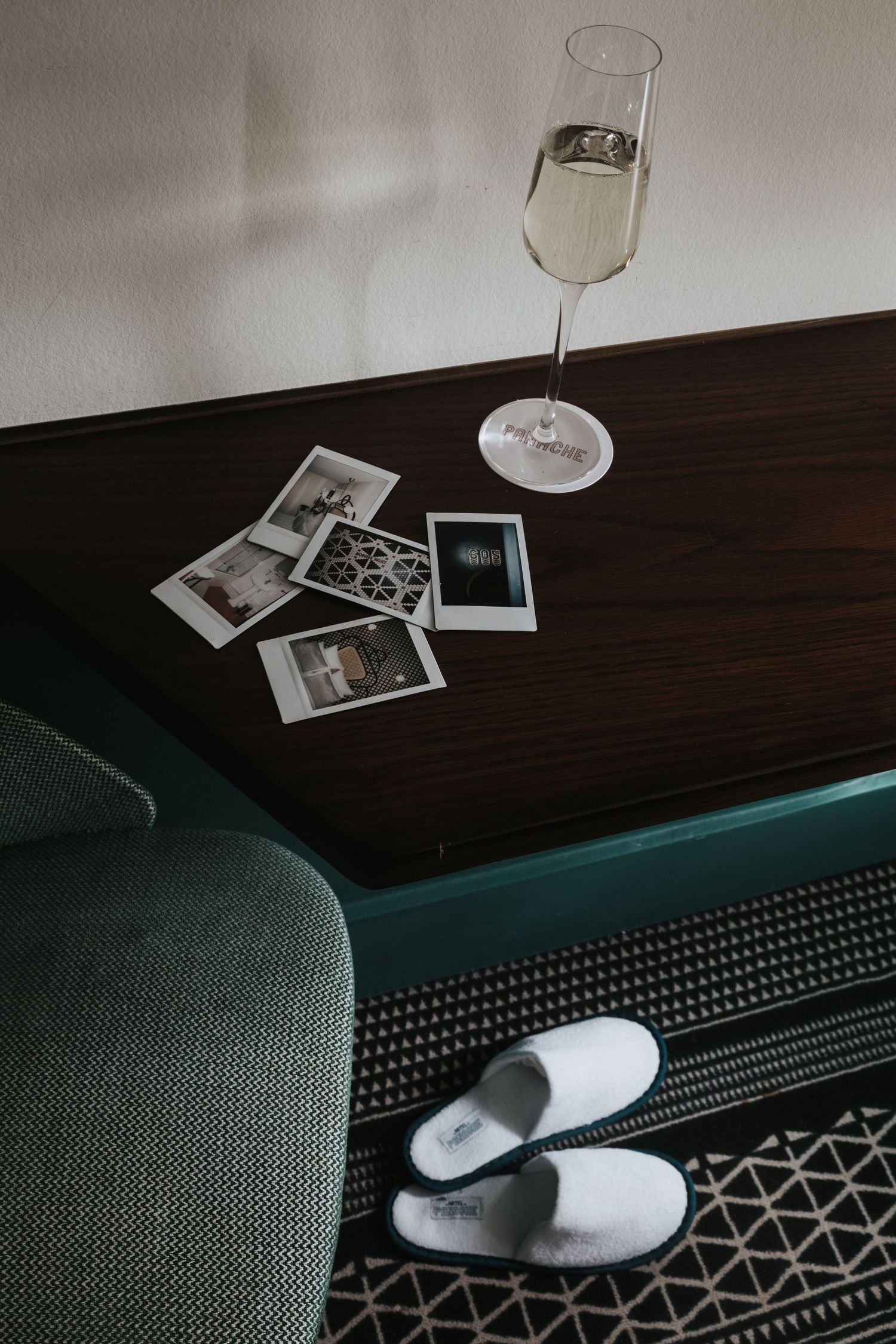 A wine glass and scattered polaroid photos rest on a dark wooden surface beside a gray couch with turquoise accent lighting.