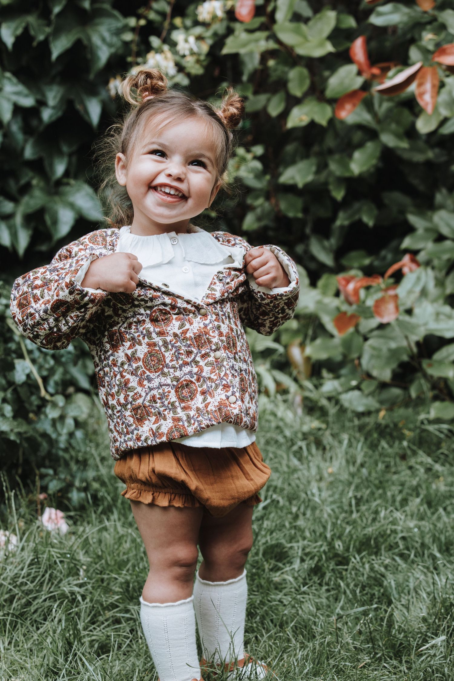 A toddler in floral dress and brown bloomers stands in a garden with a joyful expression.