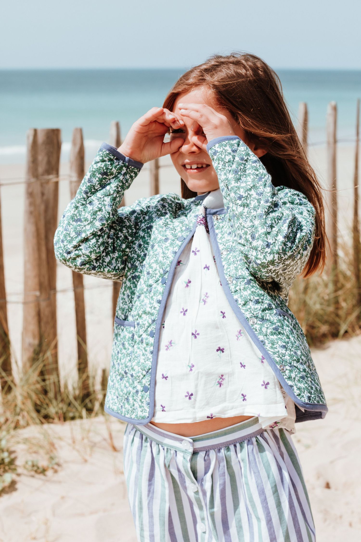 A series of photos showing someone in a floral mint green jacket and white dress posing on a sandy beach near wooden posts.