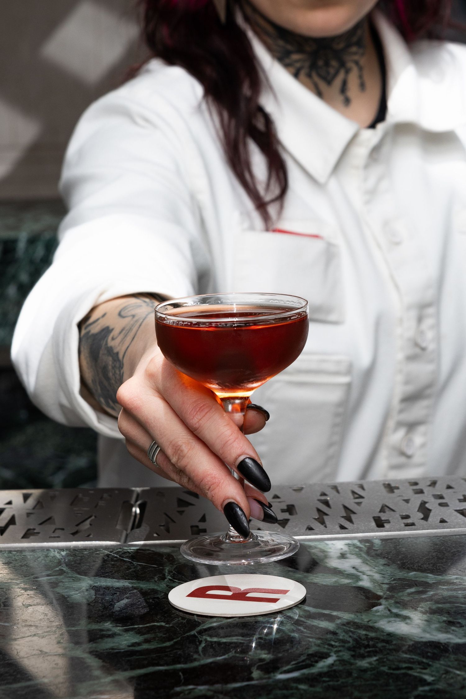 A bartender's tattooed hand holds a coupe glass with a dark cocktail against a marble bar surface.