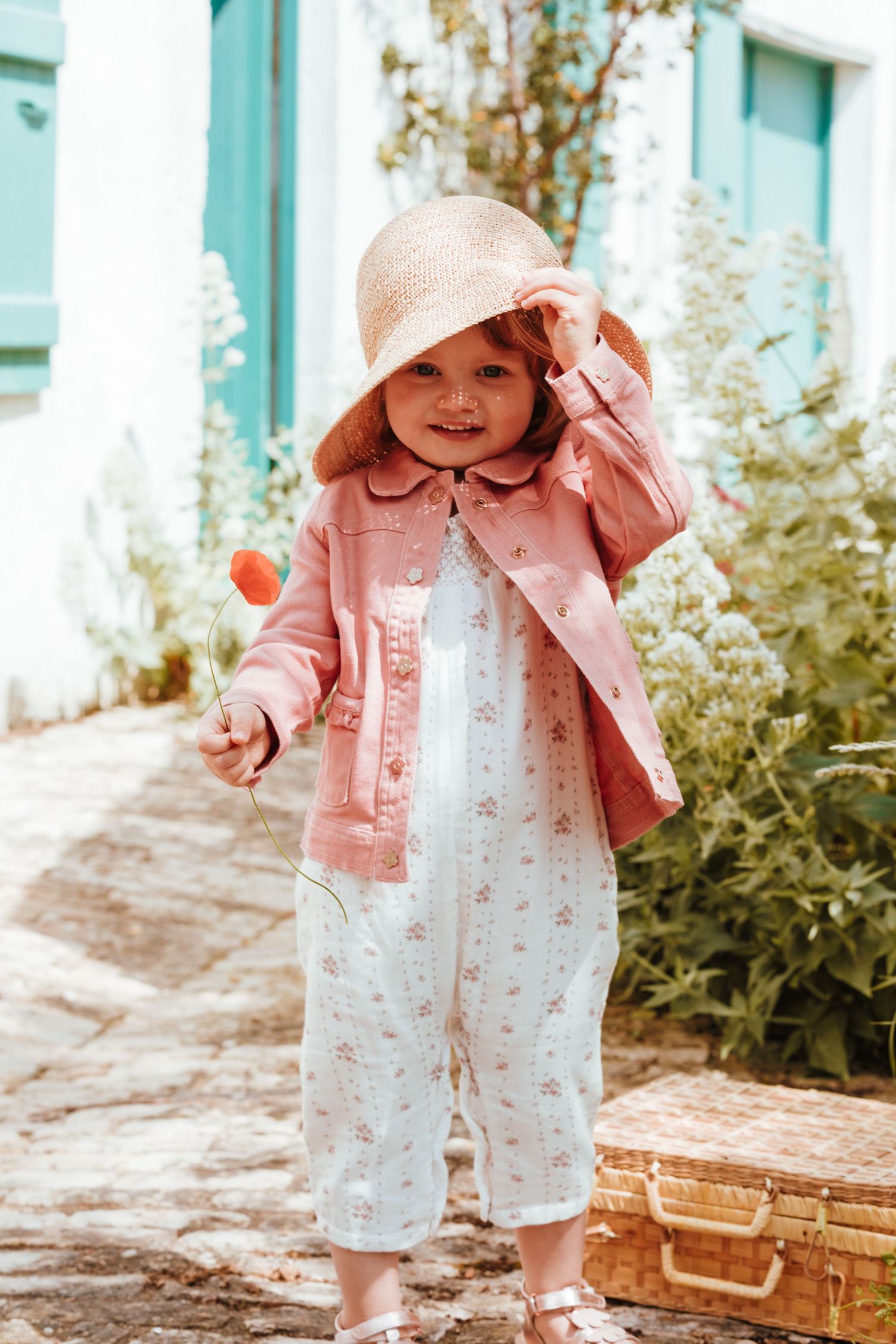 A baby wearing a pink jacket, white romper and sun hat poses next to a vintage suitcase in a garden setting.
