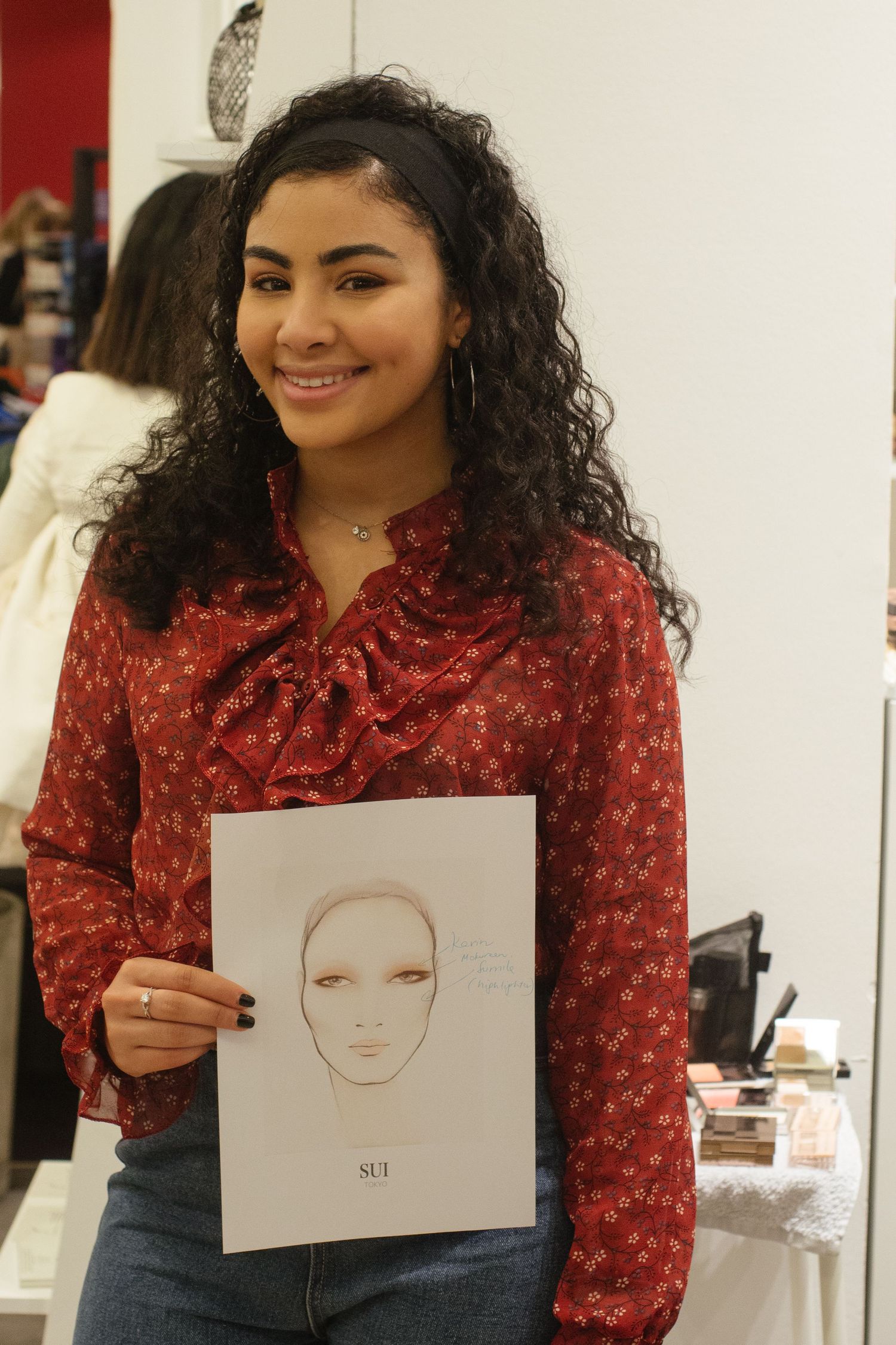 A young woman in a red patterned blouse holds up a simple face sketch while standing in a bright interior space.