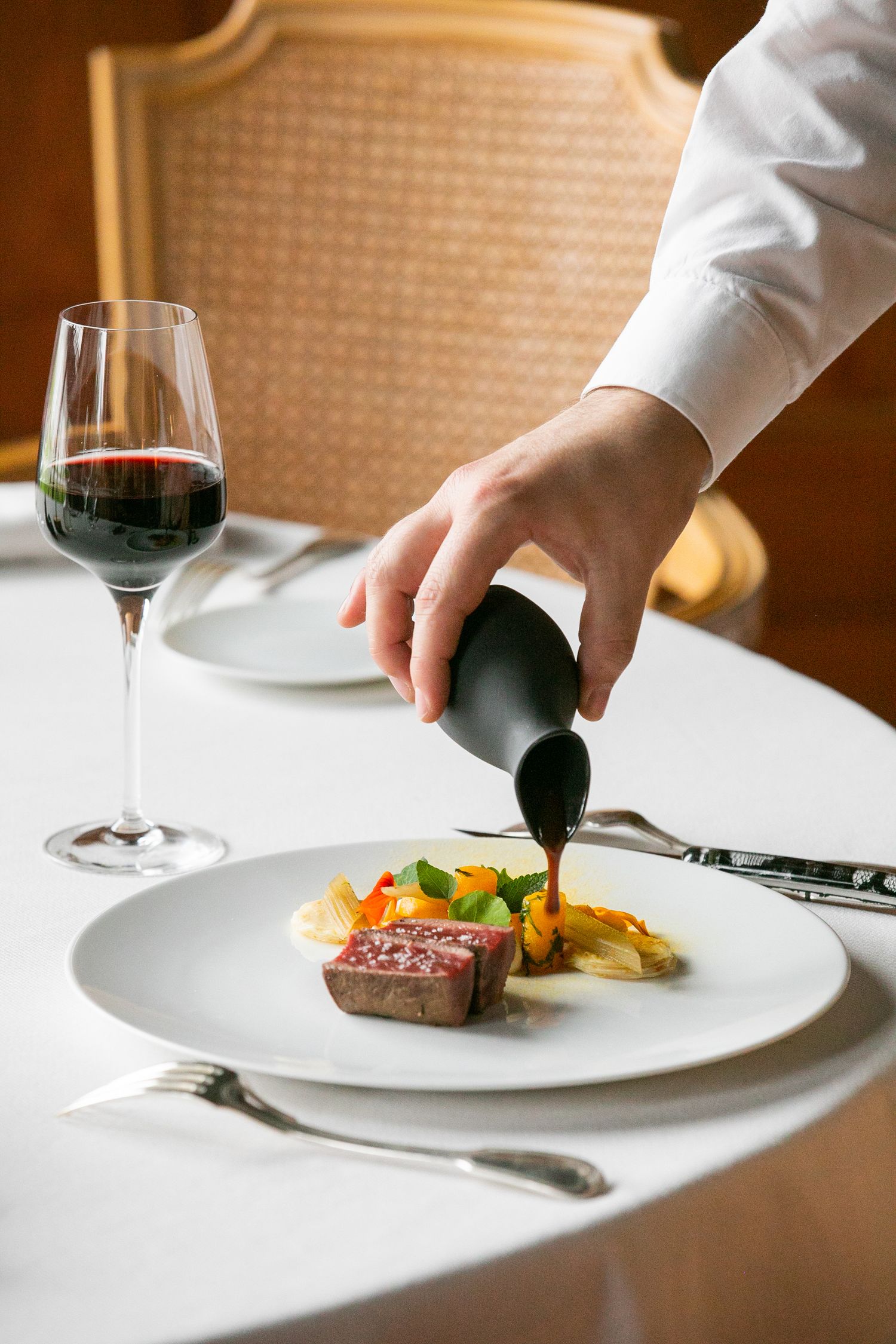 A server pours sauce over a plated steak dish with vegetables and a glass of red wine on an elegant white tablecloth.