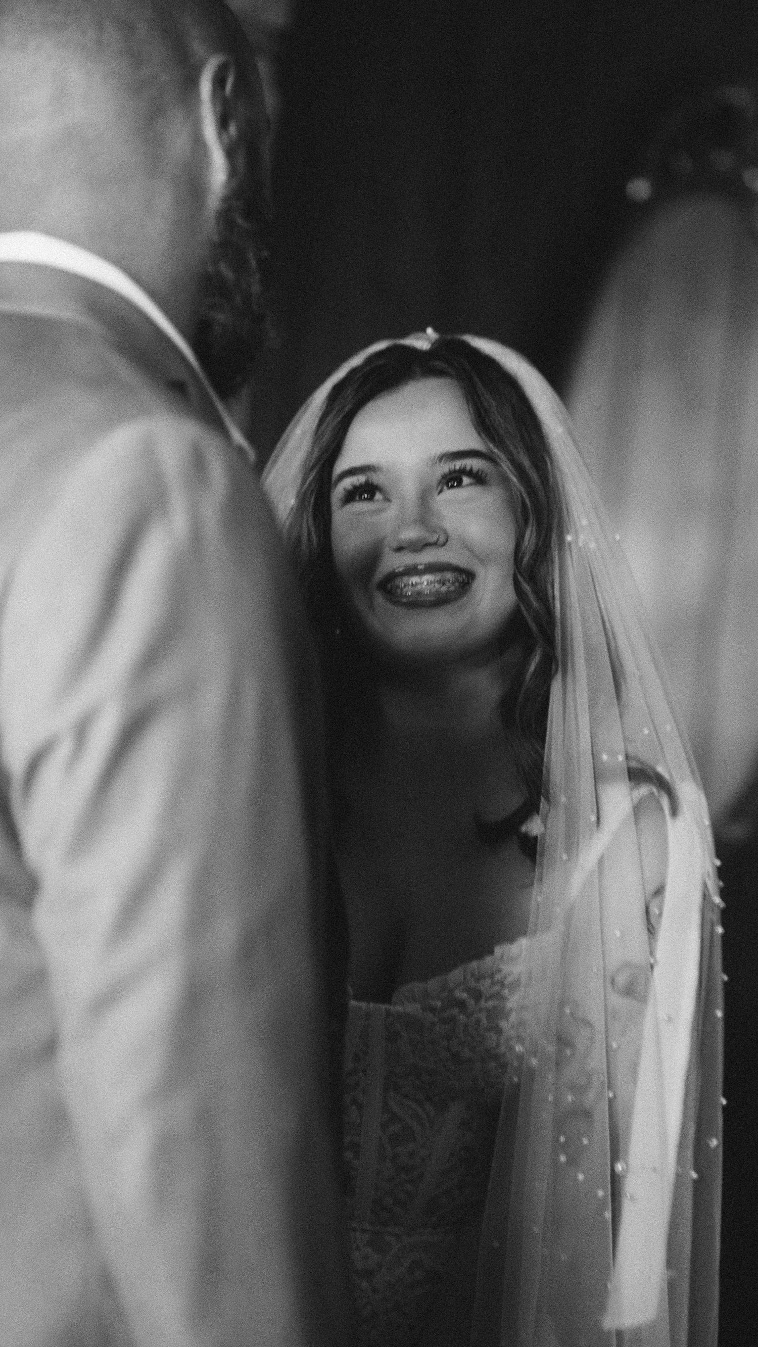 Black and white photo of a smiling bride in veil looking up at groom during wedding ceremony.