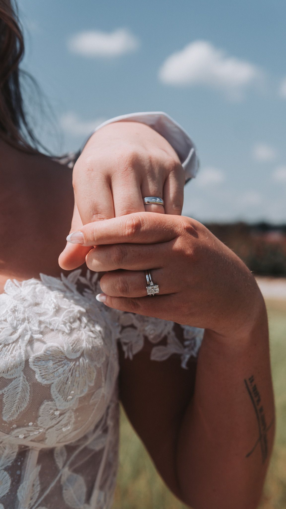 Bride and groom holding hands outdoors, showcasing diamond engagement ring and wedding band on sunny day.