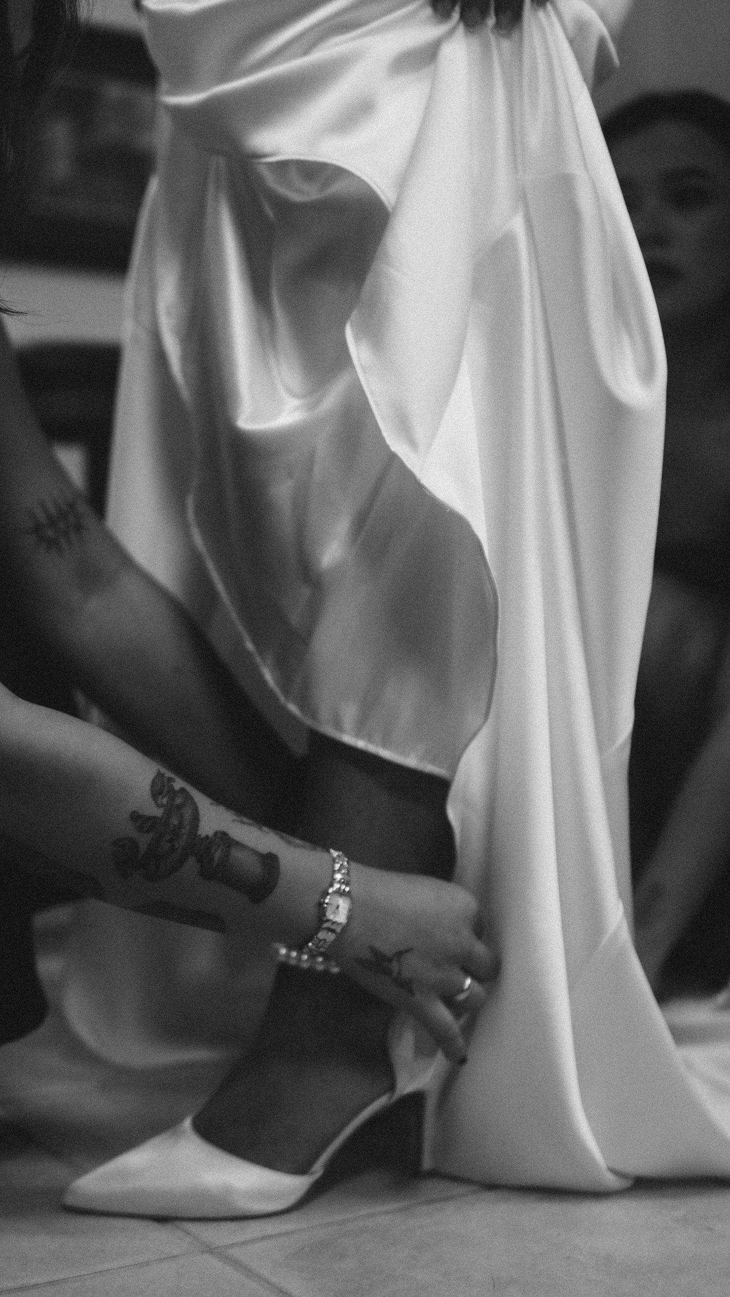 Black and white photo of tattooed hands fastening a white heeled shoe on a bride in a flowing white gown.
