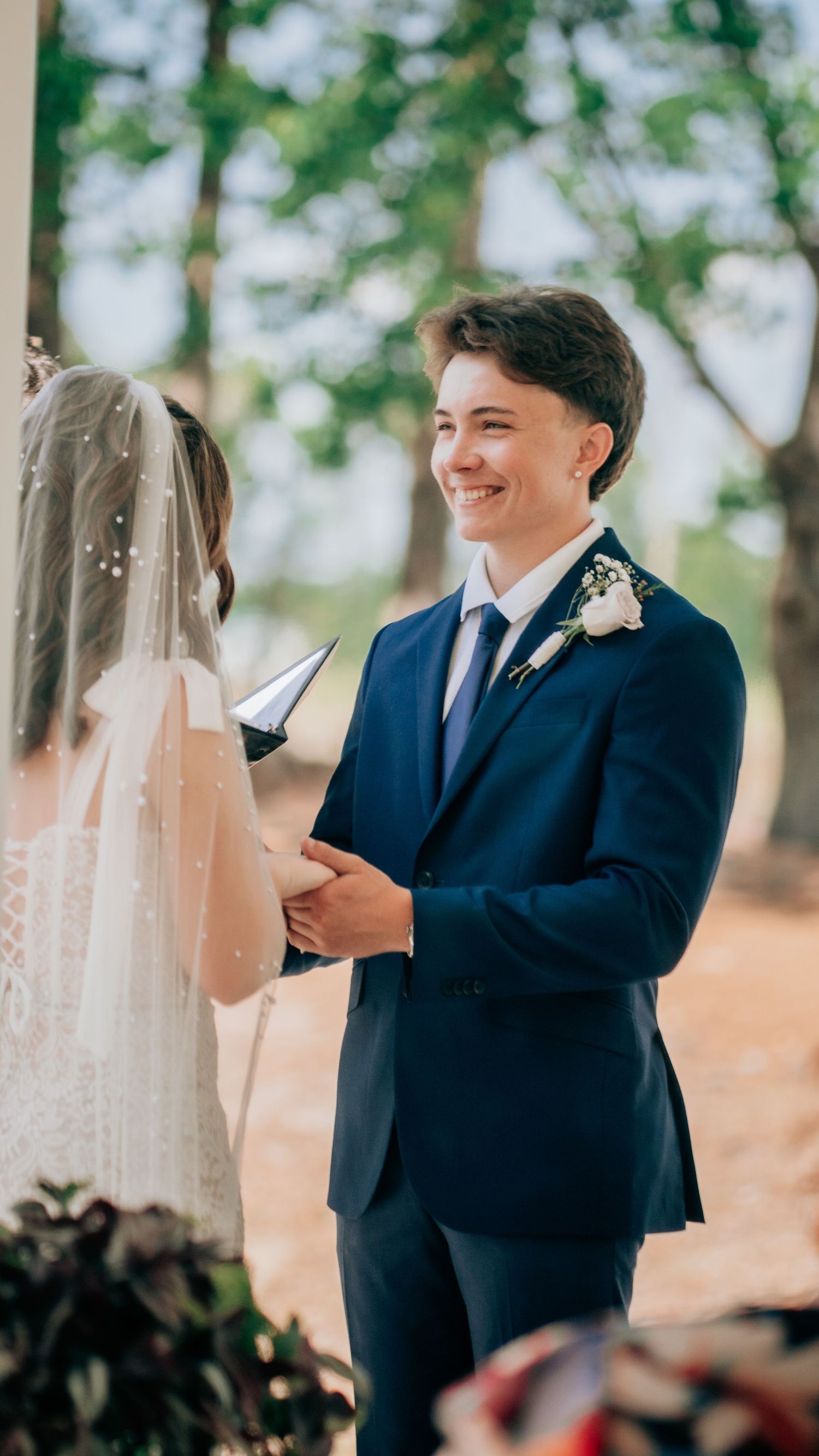 Smiling groom in navy suit with boutonniere exchanges vows with veiled bride at outdoor wedding ceremony.