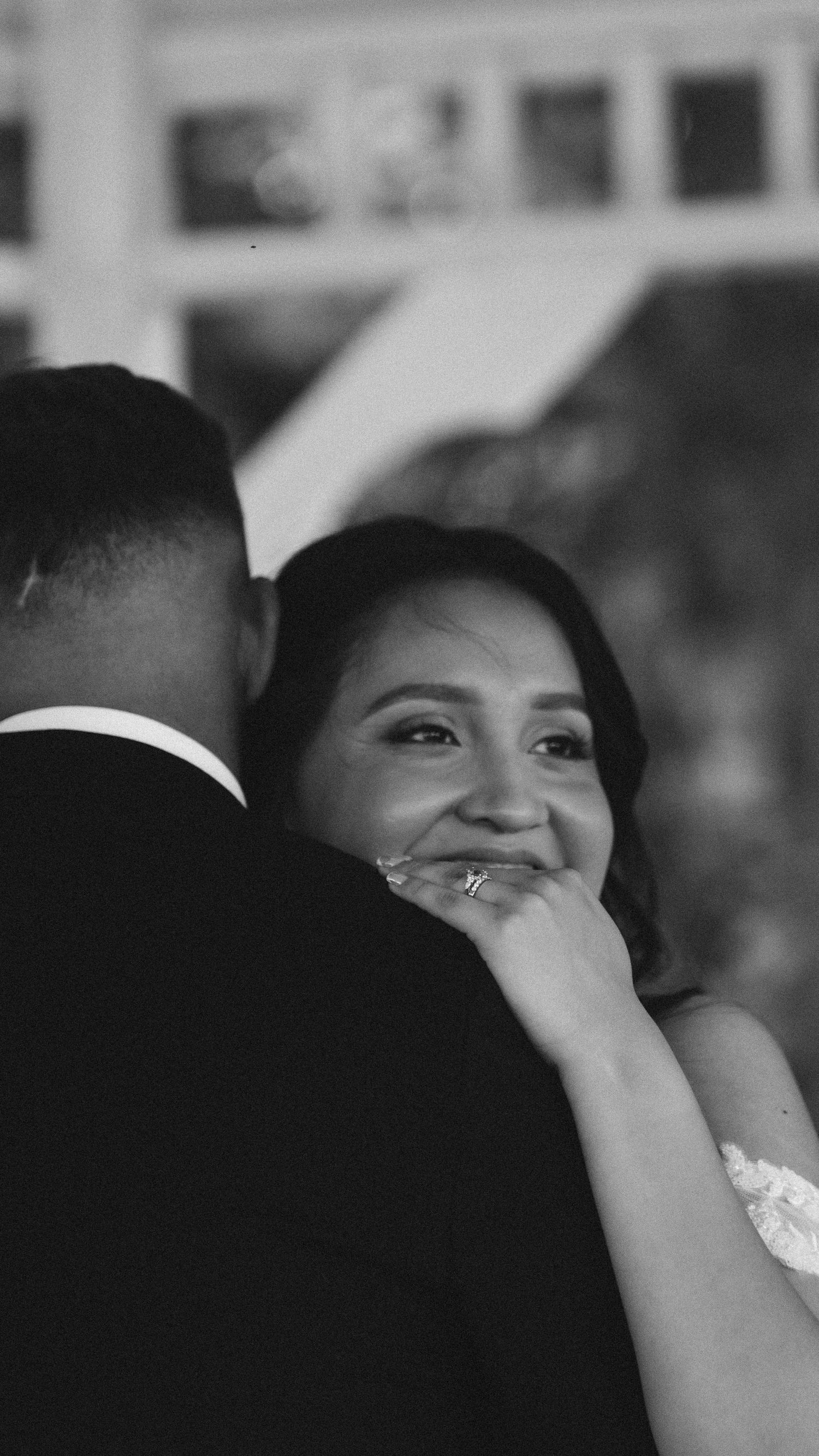 Black and white wedding photo of a smiling bride embracing her groom, wearing a lace dress and diamond ring.