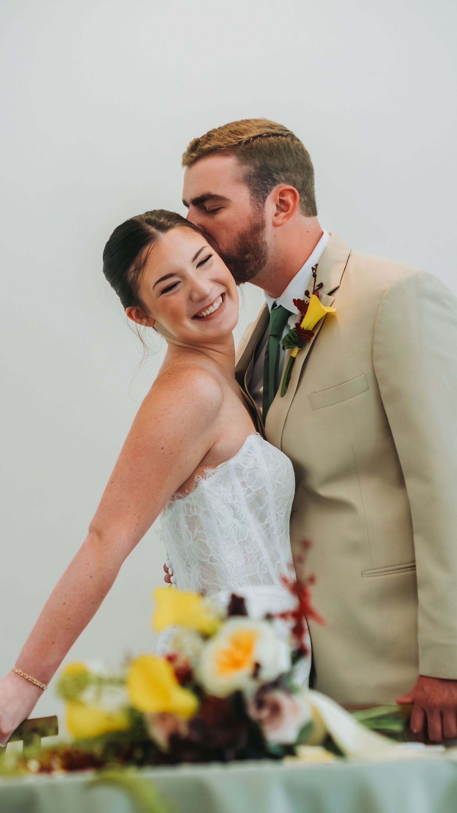 A smiling bride in a white dress receives a kiss on the cheek from a groom in a tan suit with a yellow boutonniere.