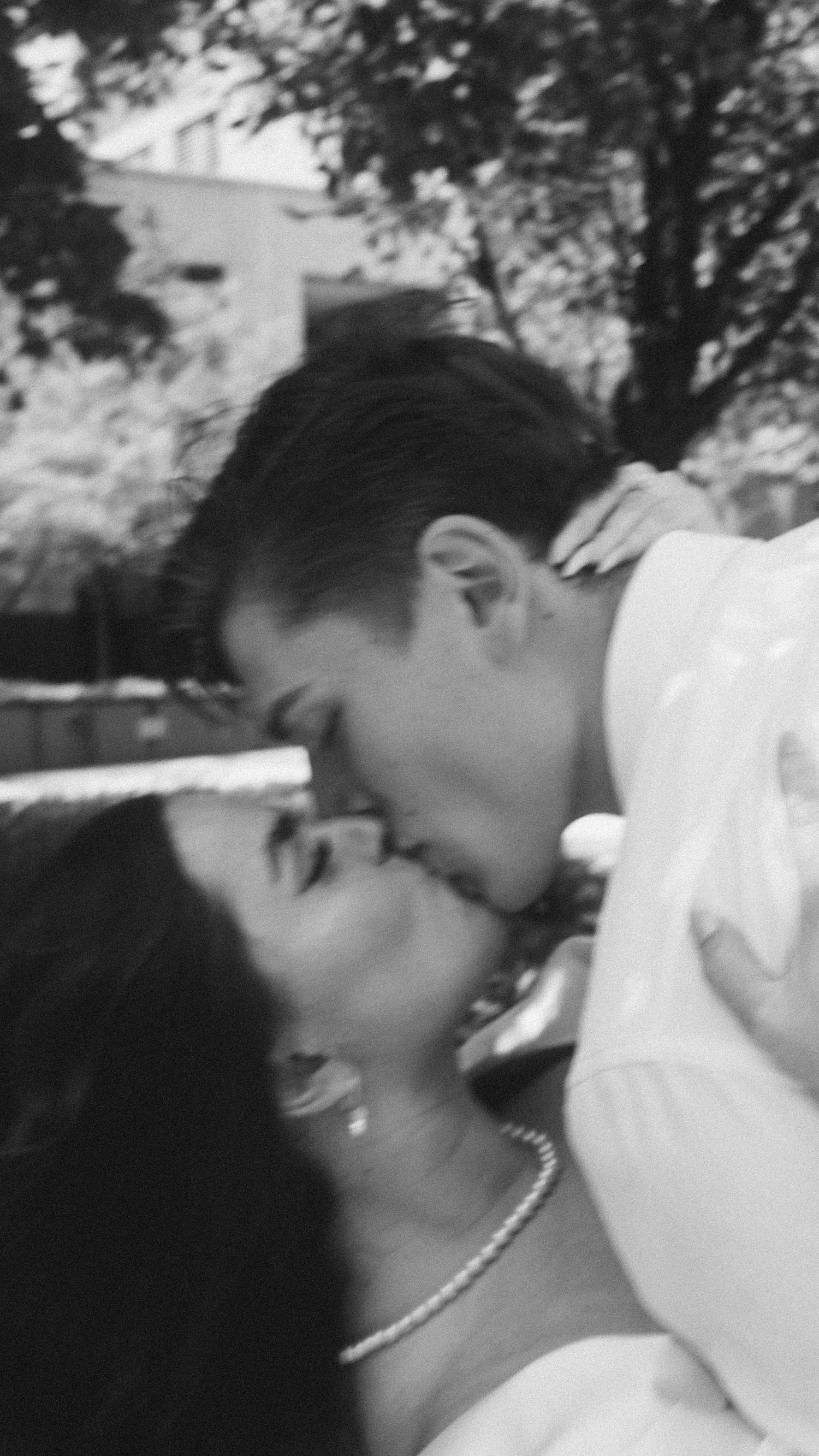 Black and white photo of a couple sharing a romantic kiss outdoors, with trees and a building in the background.
