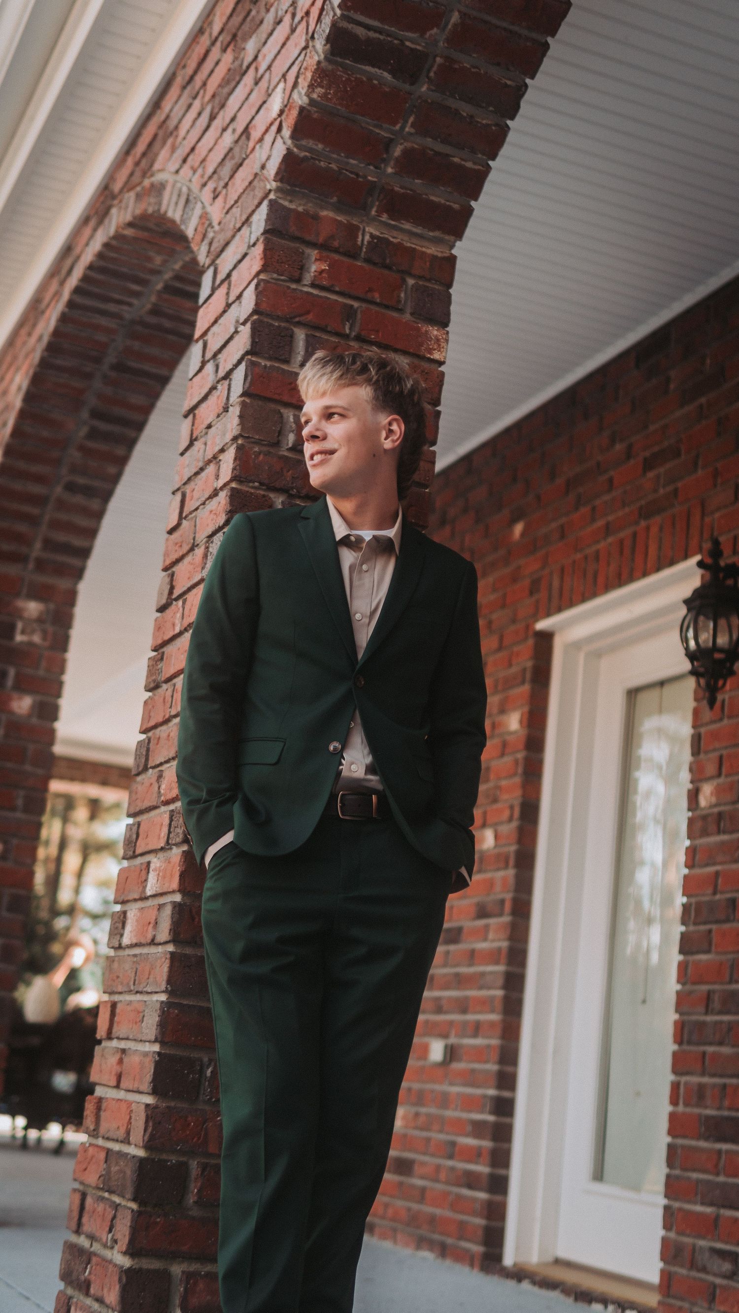 Stylish young man in a dark green suit standing confidently under brick arches.