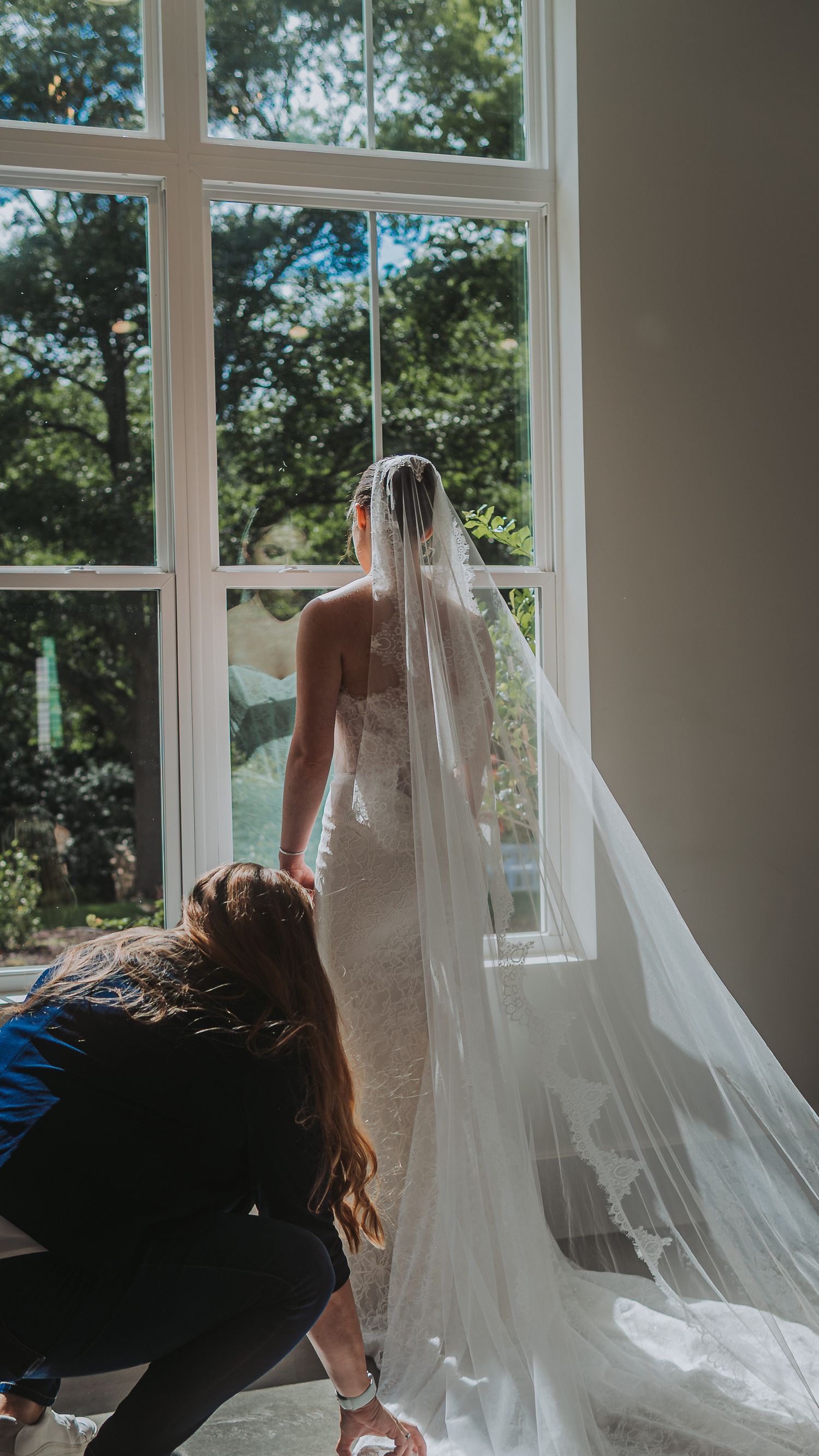 A bride in a lace gown and long veil stands by a sunlit window while an assistant adjusts her dress.
