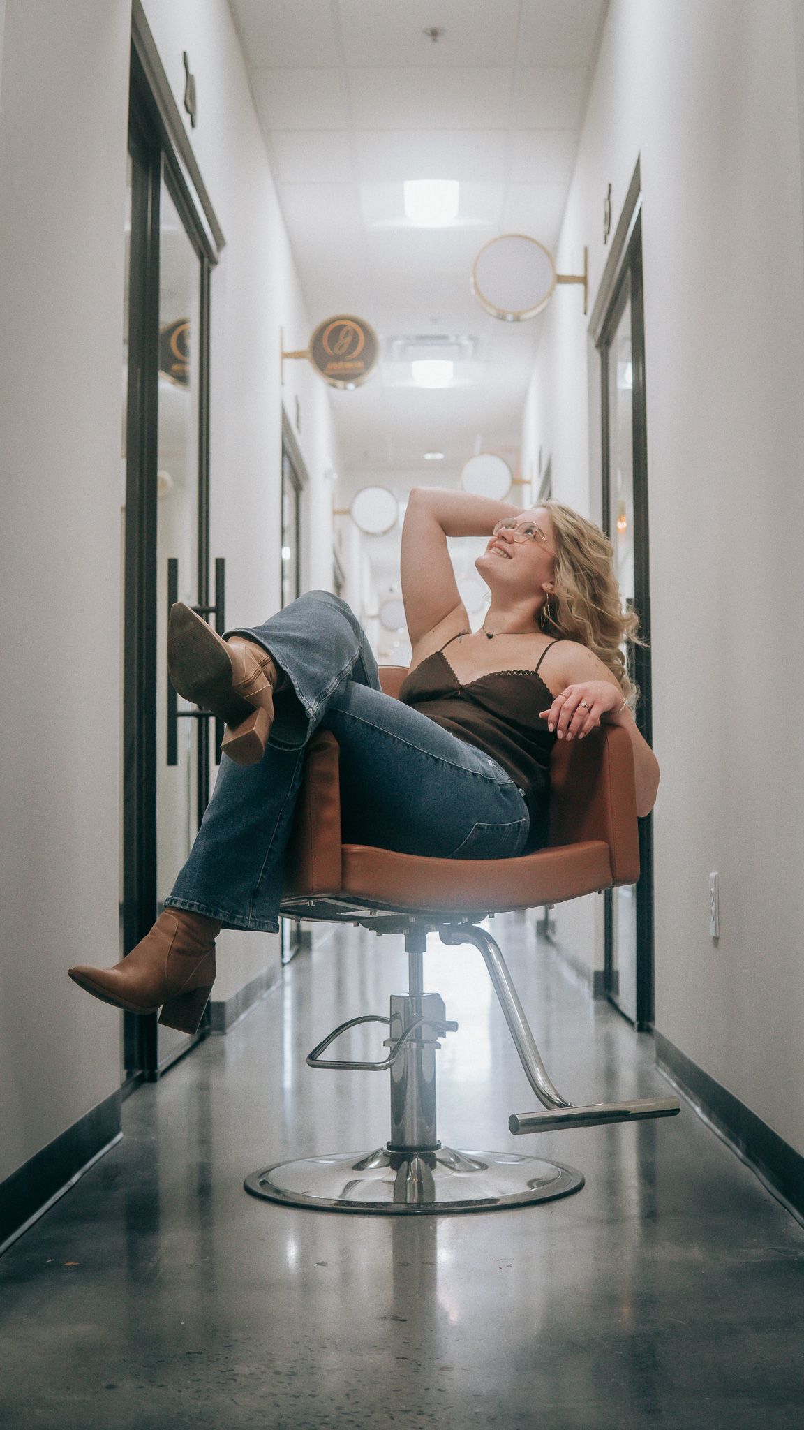 Woman relaxing in a salon chair in a hallway, wearing jeans and boots, smiling upward in a modern salon.