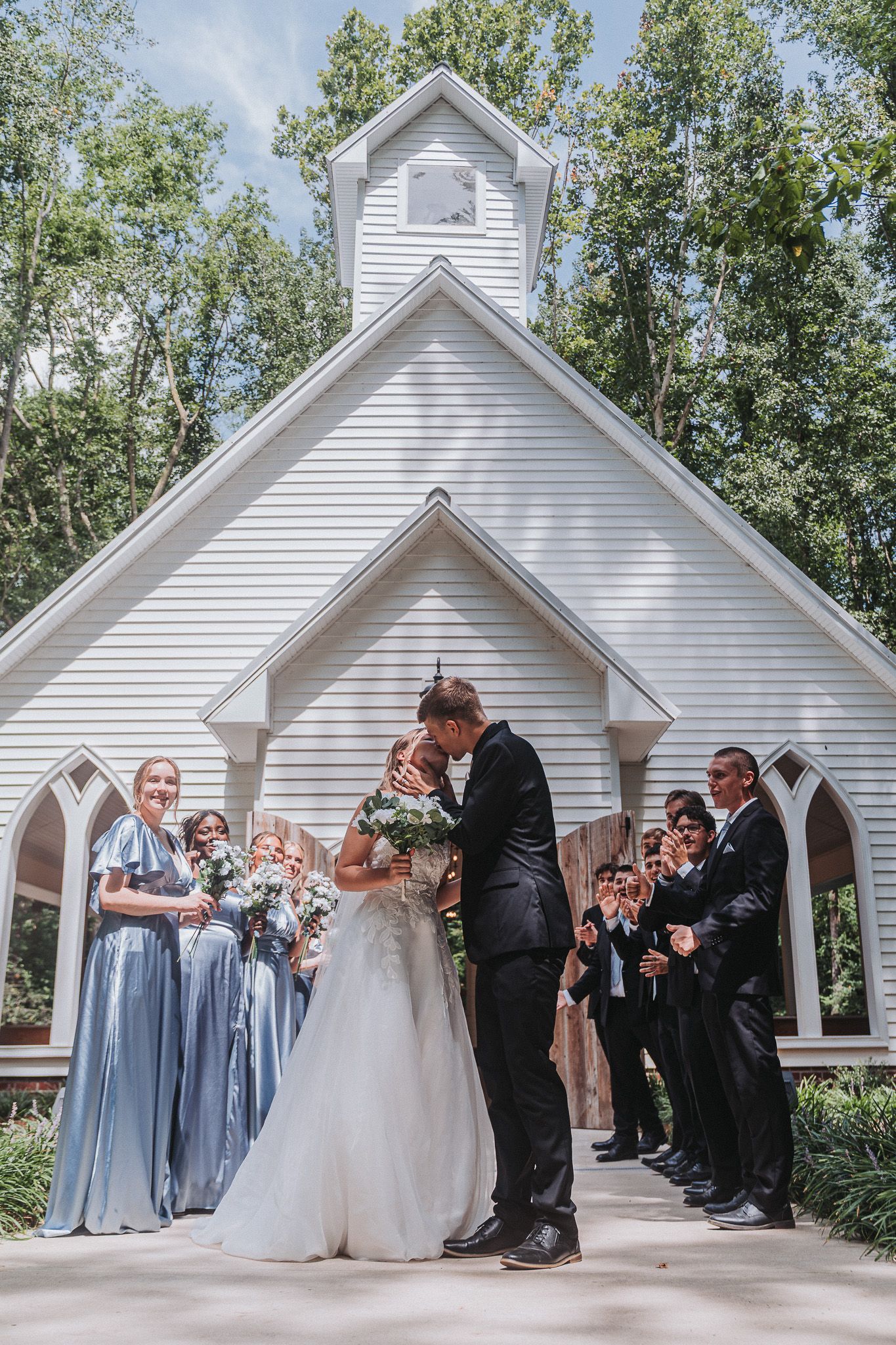 Bride and groom share first kiss outside a white chapel surrounded by bridesmaids in blue and groomsmen.