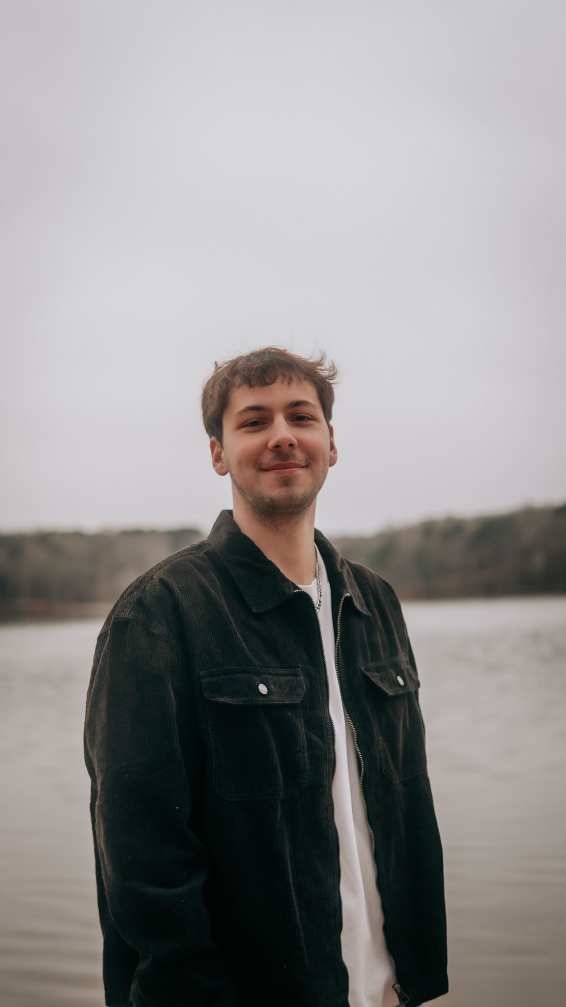 Young man smiling in black jacket standing near a lake on an overcast day.