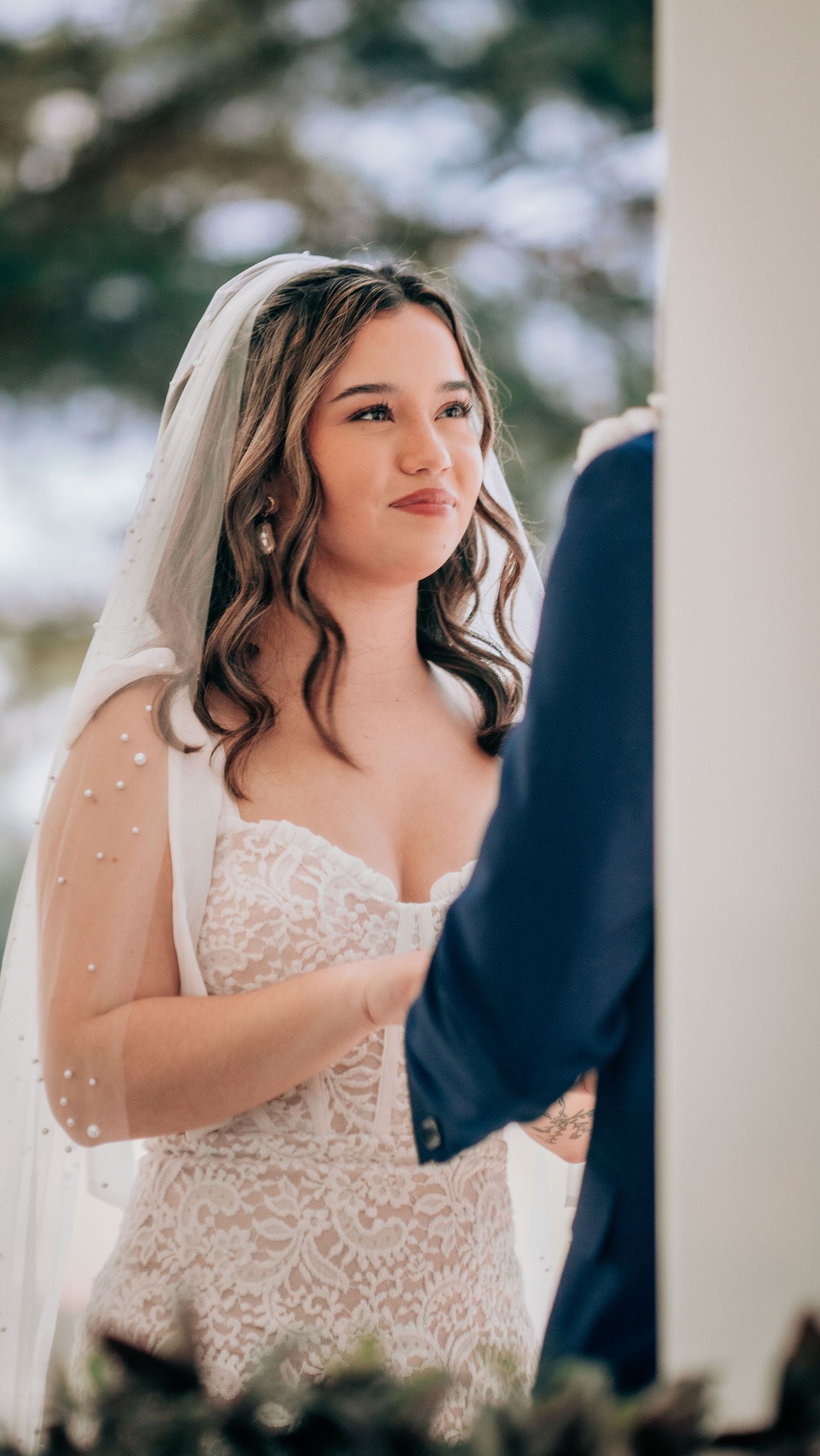 Bride in white lace gown and pearl-trimmed veil smiles during outdoor wedding ceremony.