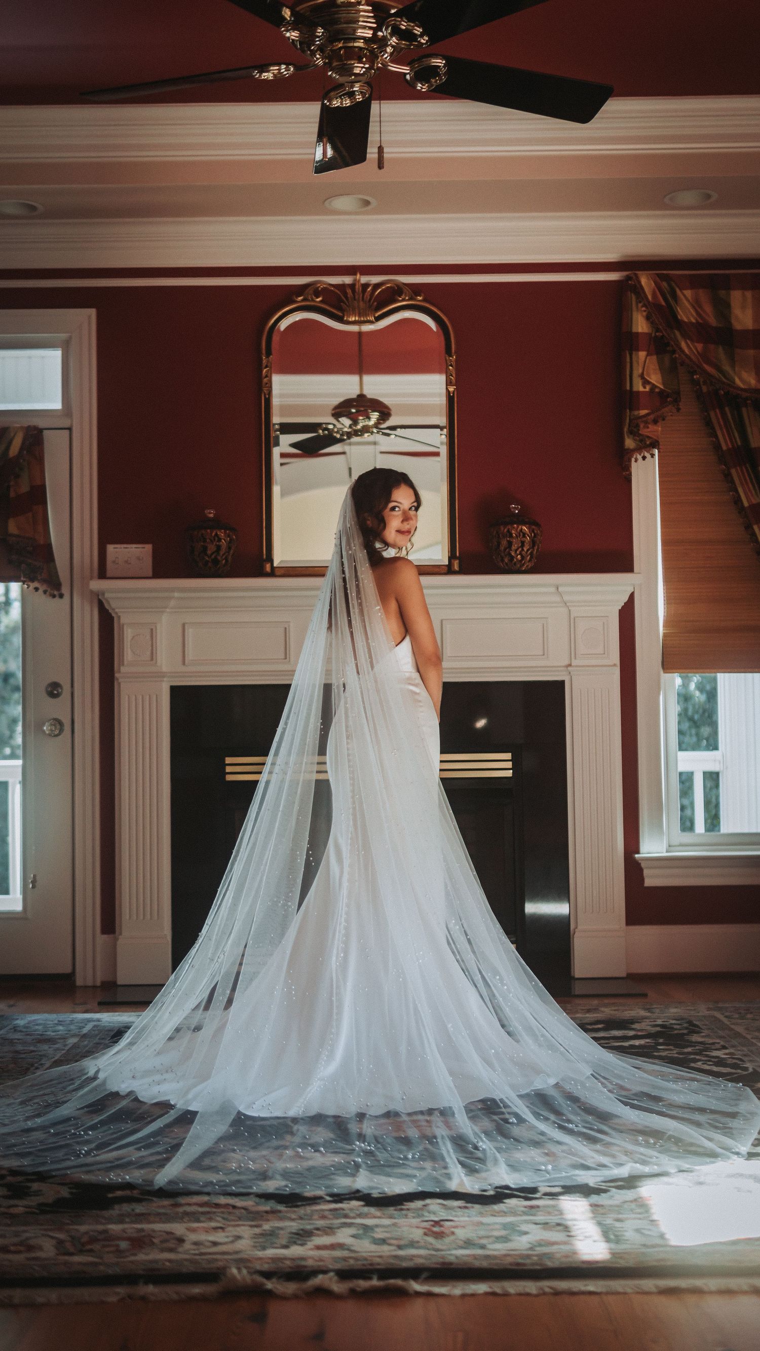 Bride in elegant white wedding gown with long flowing veil stands before fireplace in red-walled room.