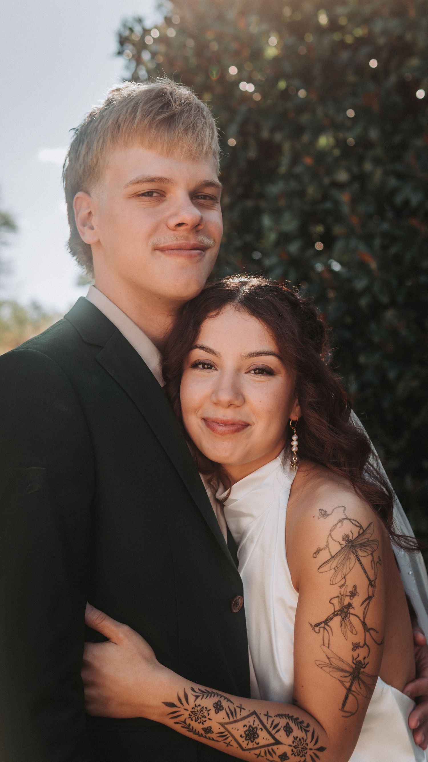 A young couple in wedding attire smiles warmly outdoors, the bride showing elegant arm tattoos in soft golden sunlight.