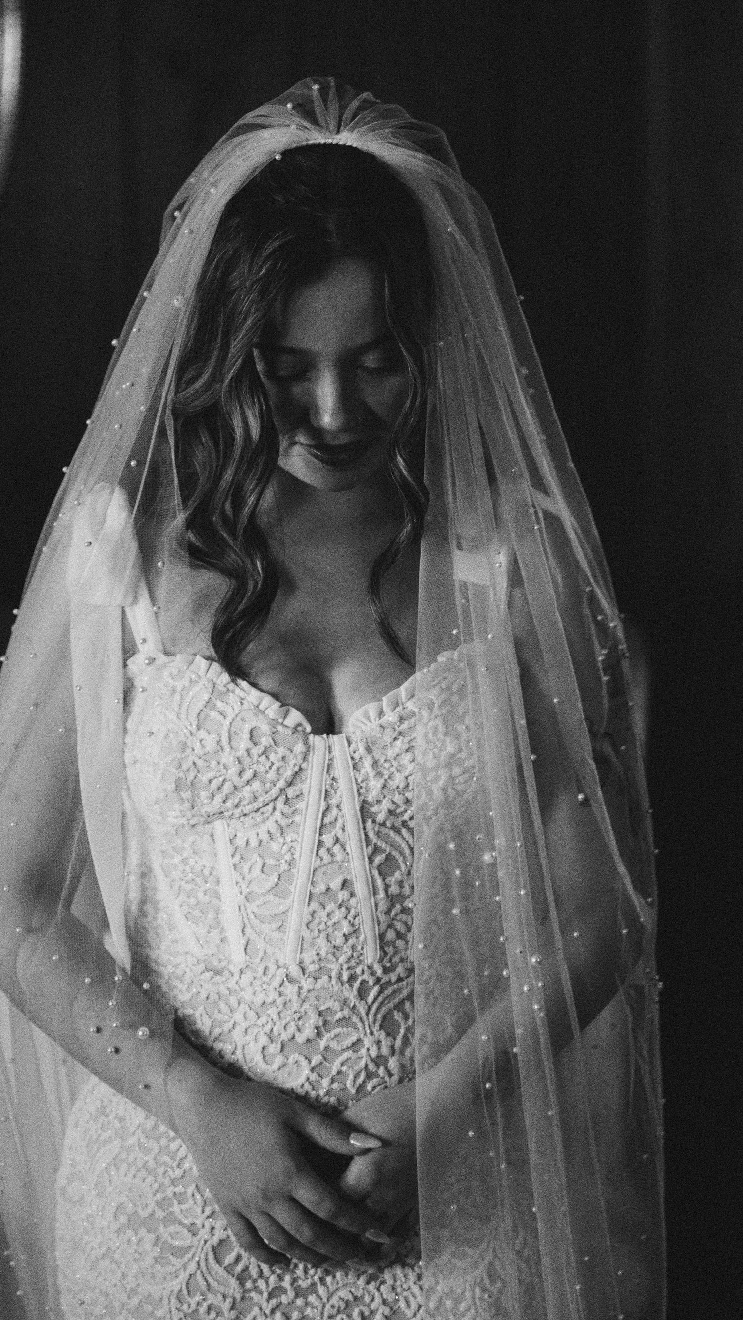 Bride in lace gown and pearl-adorned veil smiles softly downward in elegant black and white wedding portrait.