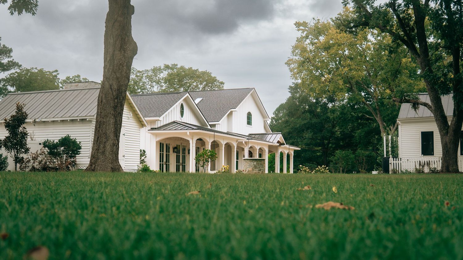 White farmhouse with covered porch surrounded by lush green lawn and mature trees on a cloudy day.