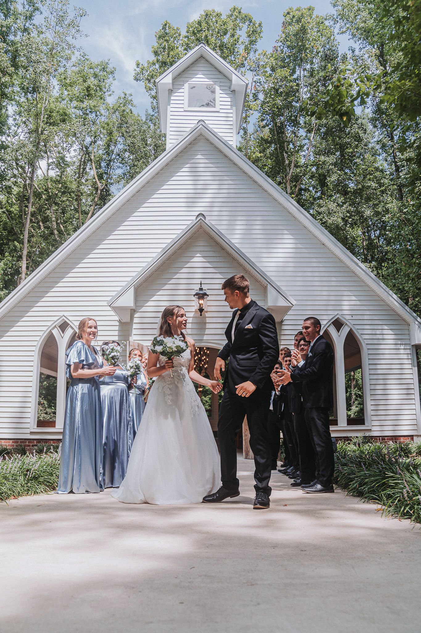 Bride and groom hold hands outside a white chapel surrounded by bridesmaids in blue and groomsmen in black suits.