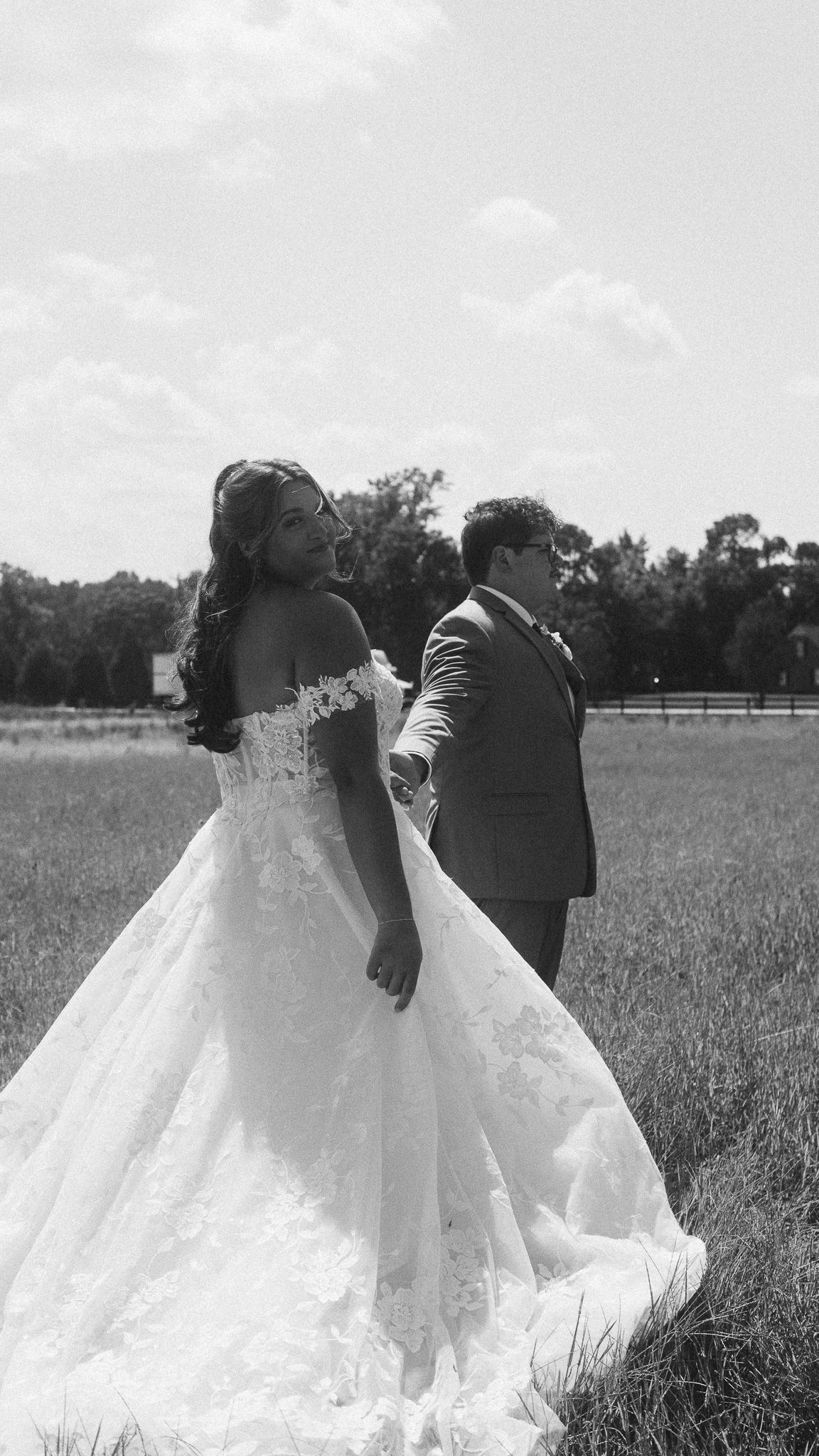 Black and white photo of bride in ball gown and groom holding hands in open field.