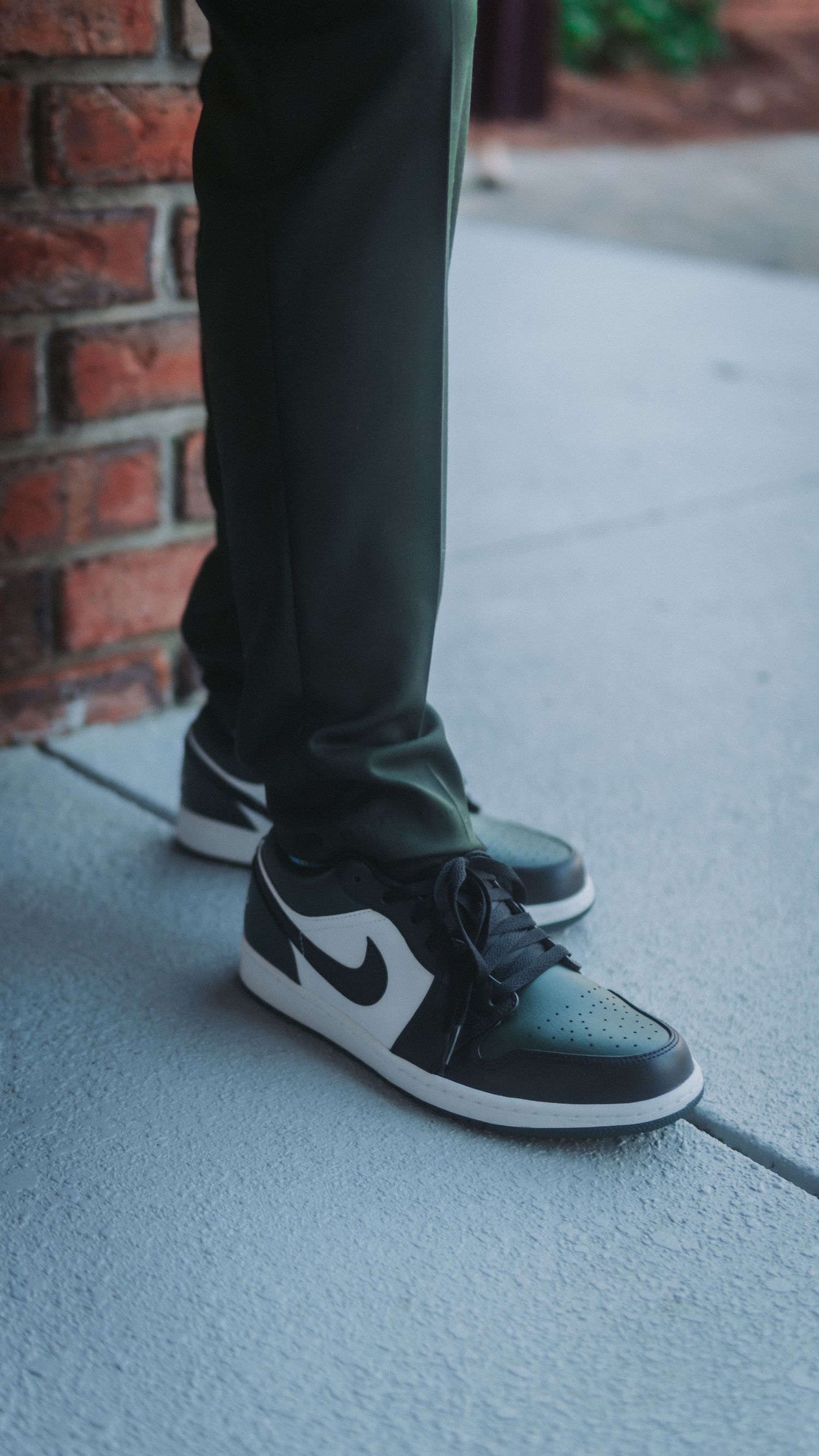 Person wearing olive green pants and black and white Nike Air Jordan 1 Low sneakers standing on concrete near brick wall.