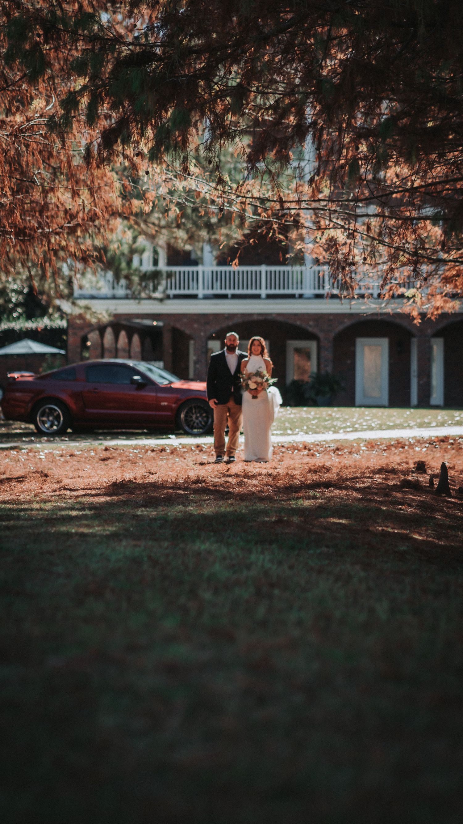 Bride and groom standing together outdoors near a red car and historic home surrounded by autumn trees.