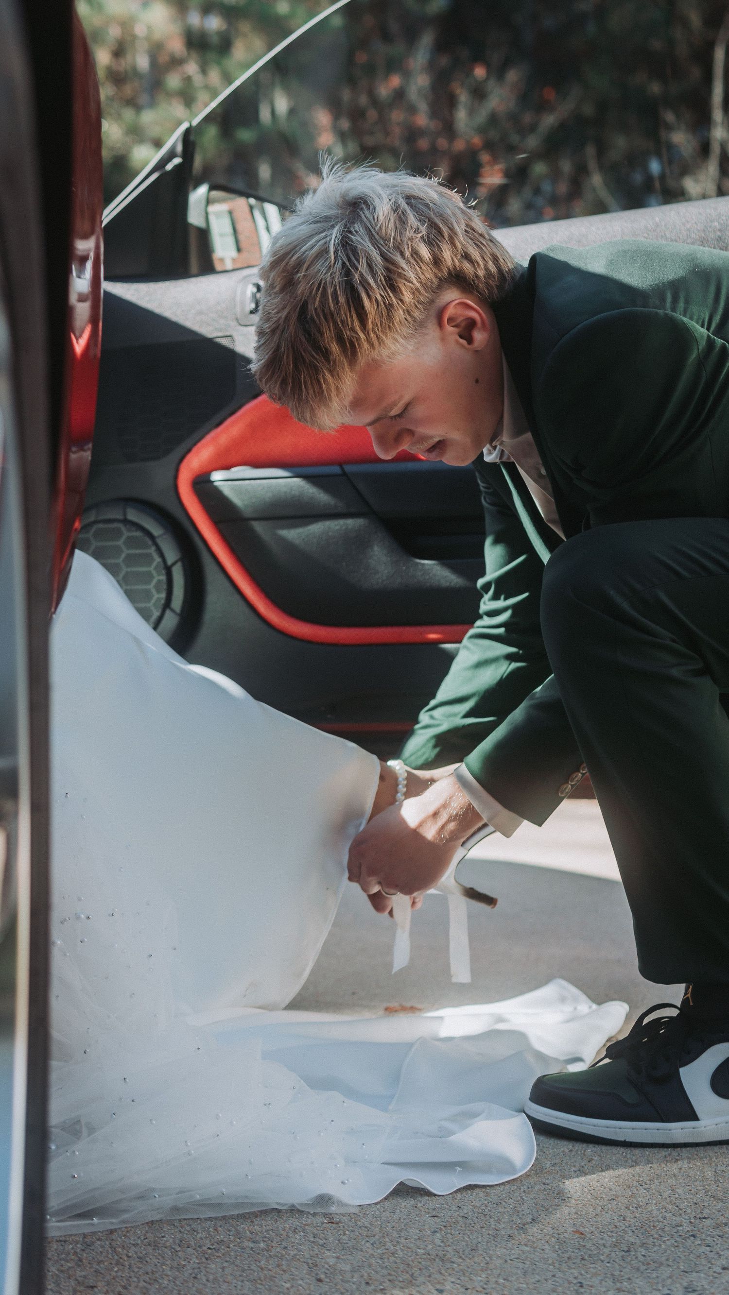 Groom in green suit carefully arranges bride's white wedding dress train beside a red sports car.