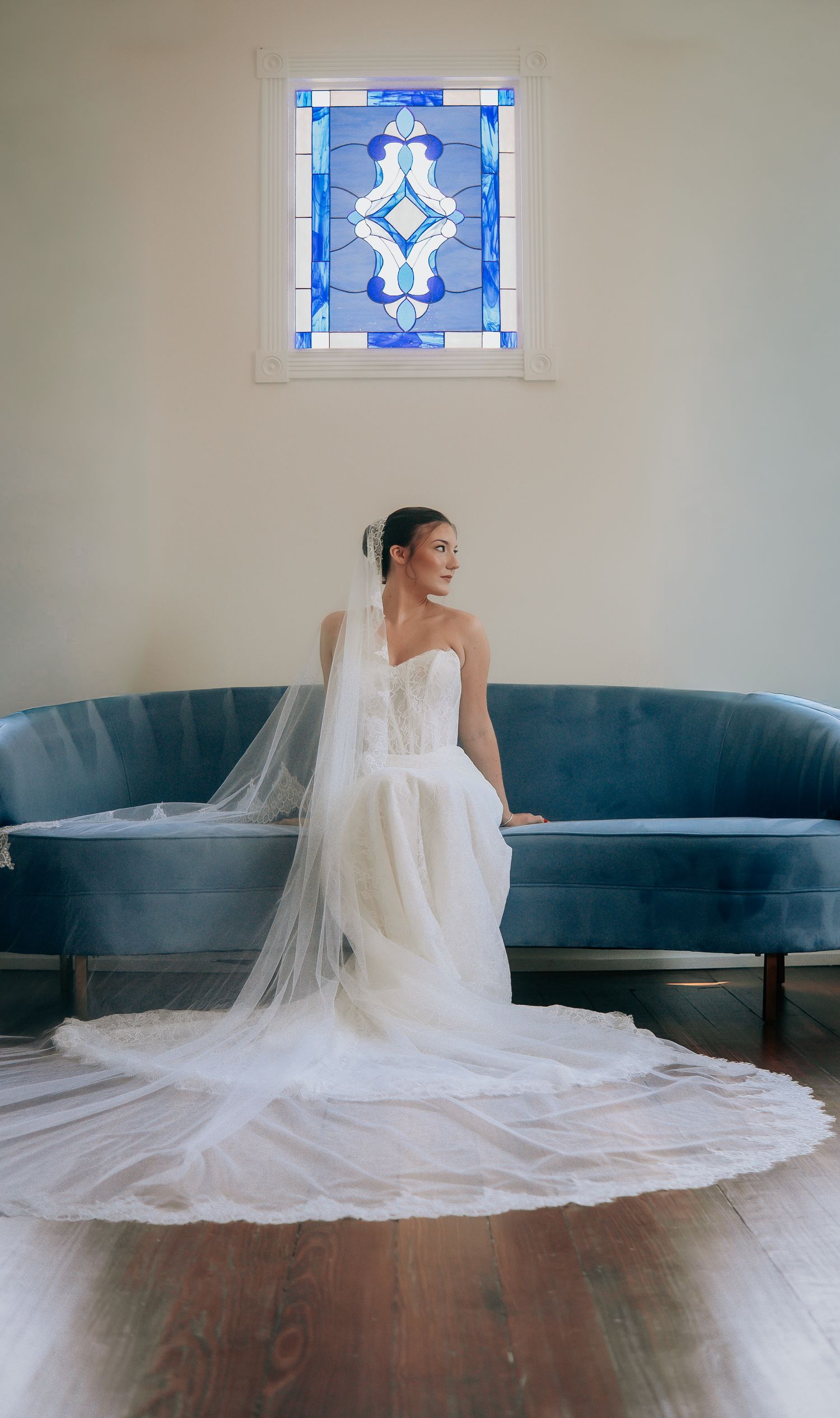 Bride in a strapless white gown with long veil sits before a blue velvet sofa beneath a stained glass window.