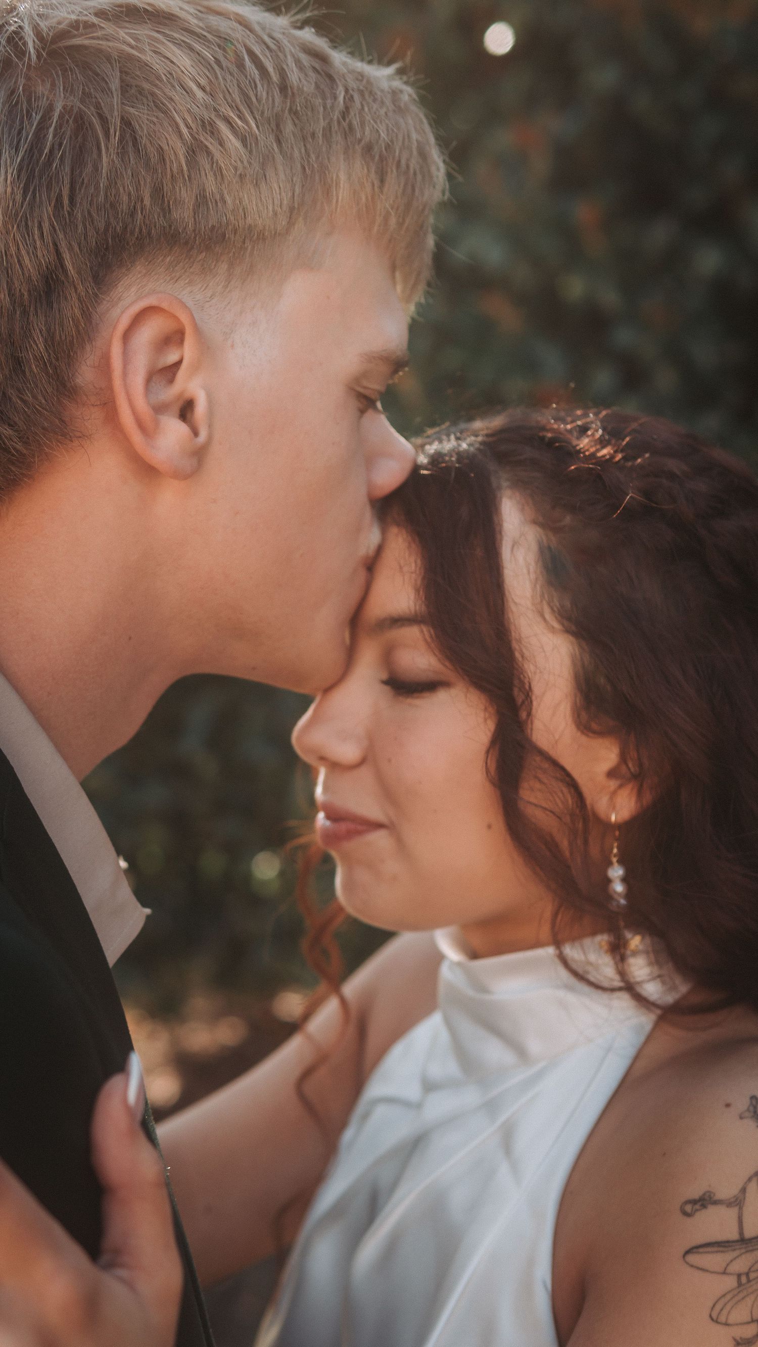 A groom tenderly kisses his bride on the forehead during a romantic outdoor wedding portrait session.