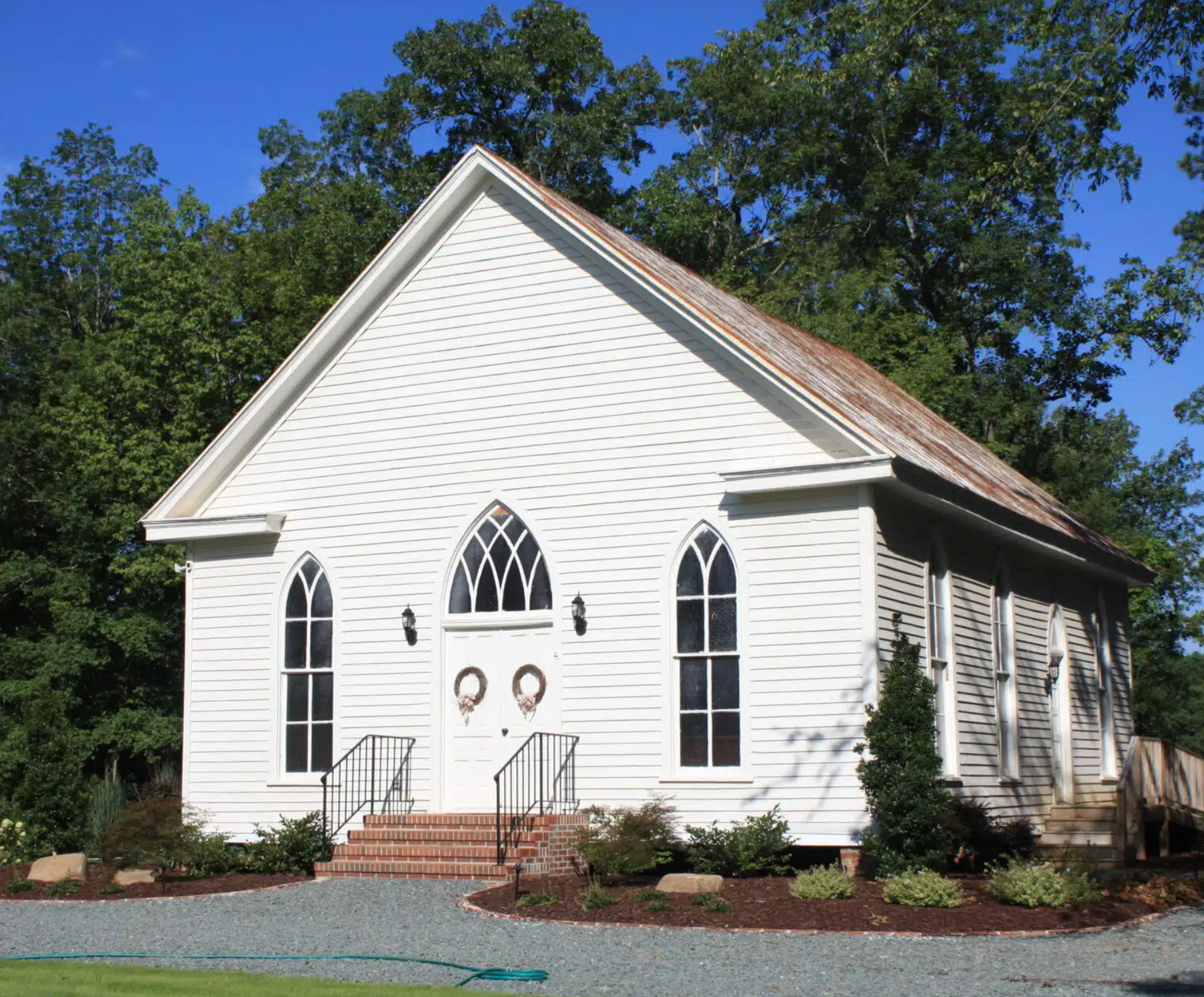 A charming white wooden church with Gothic arched windows, brick steps, and lush green trees under a clear blue sky.