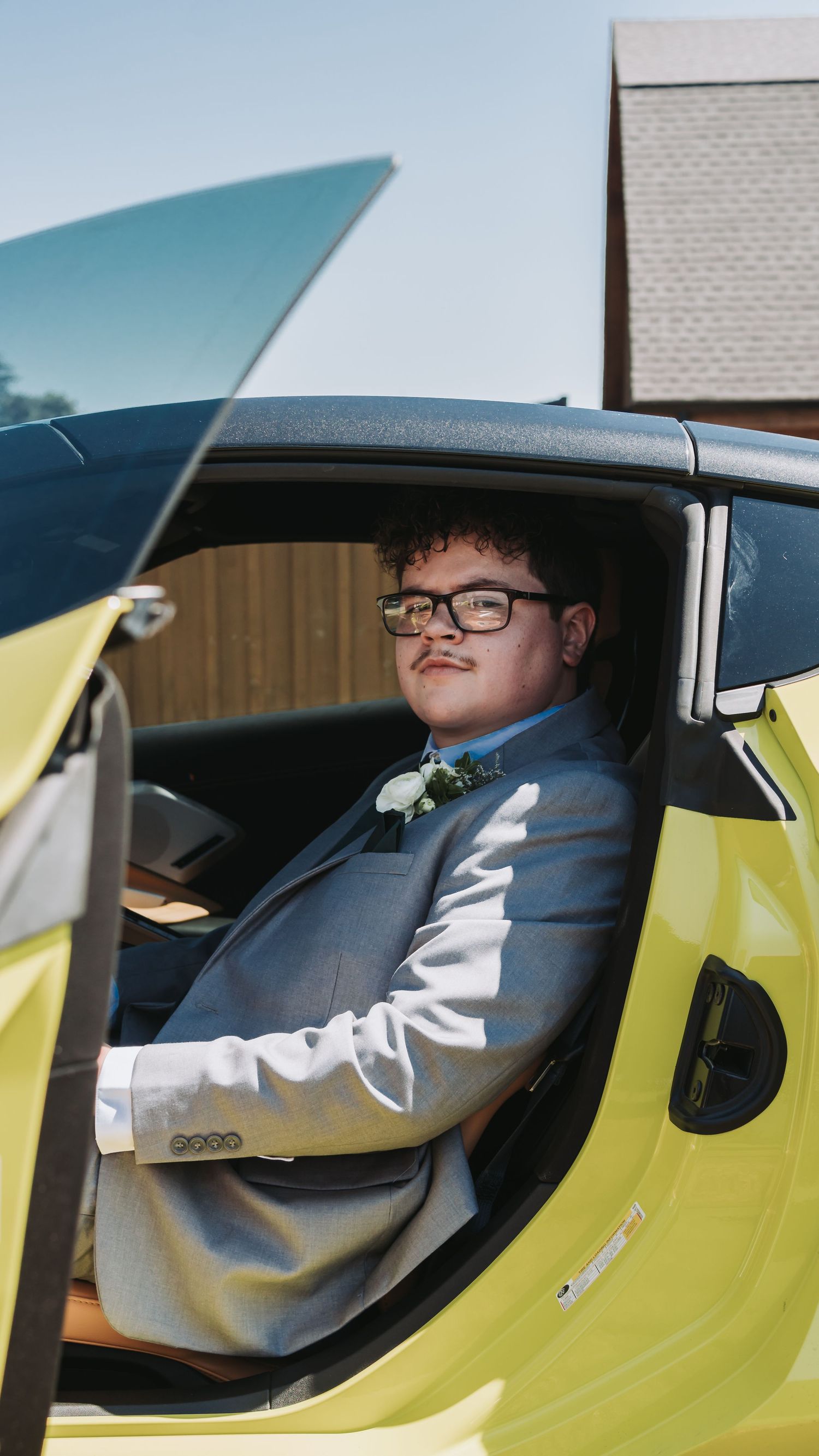 Young man in gray suit with boutonniere sits in yellow sports car at a formal occasion.