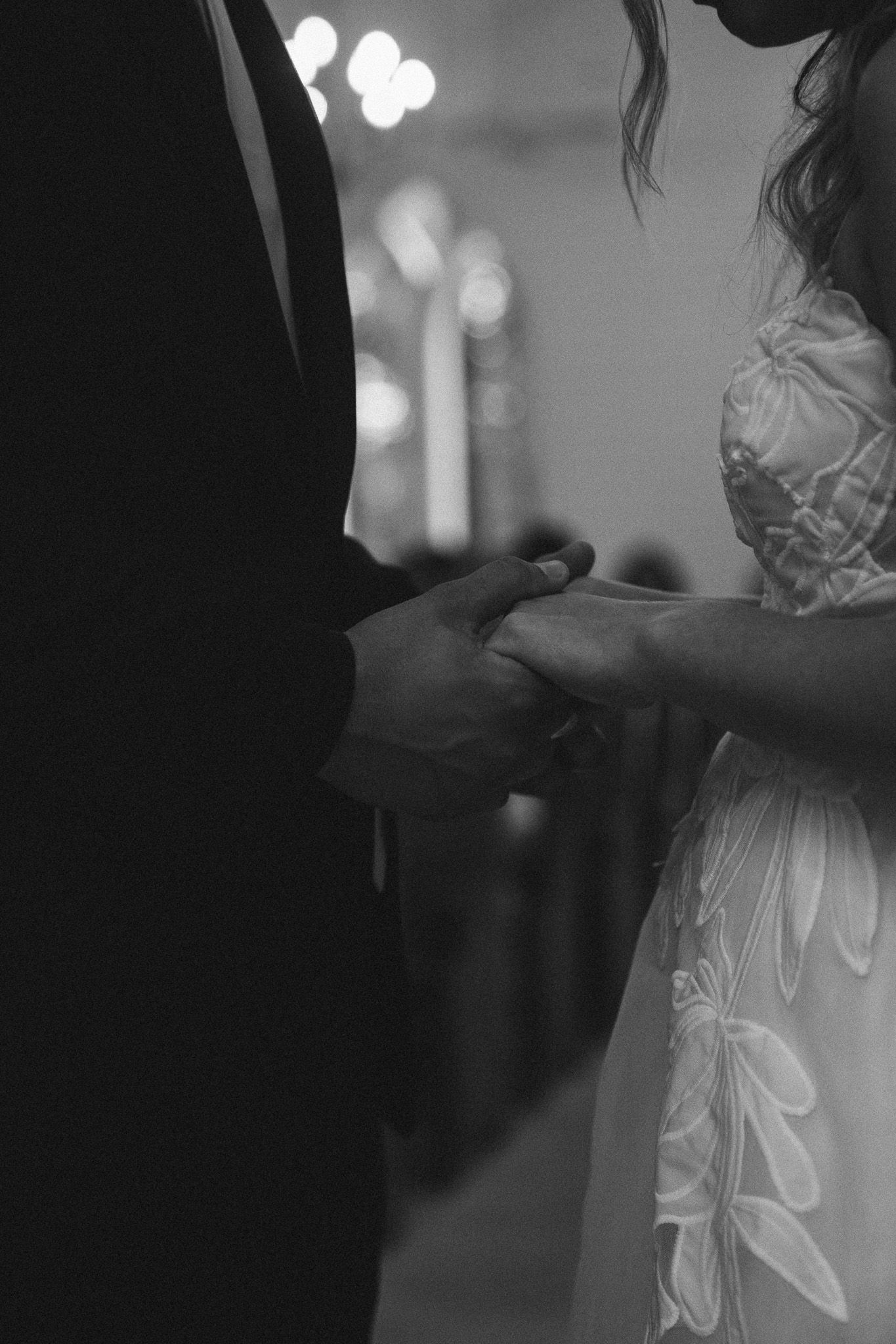 Black and white wedding photo of bride and groom holding hands during ceremony with bokeh lights in background.