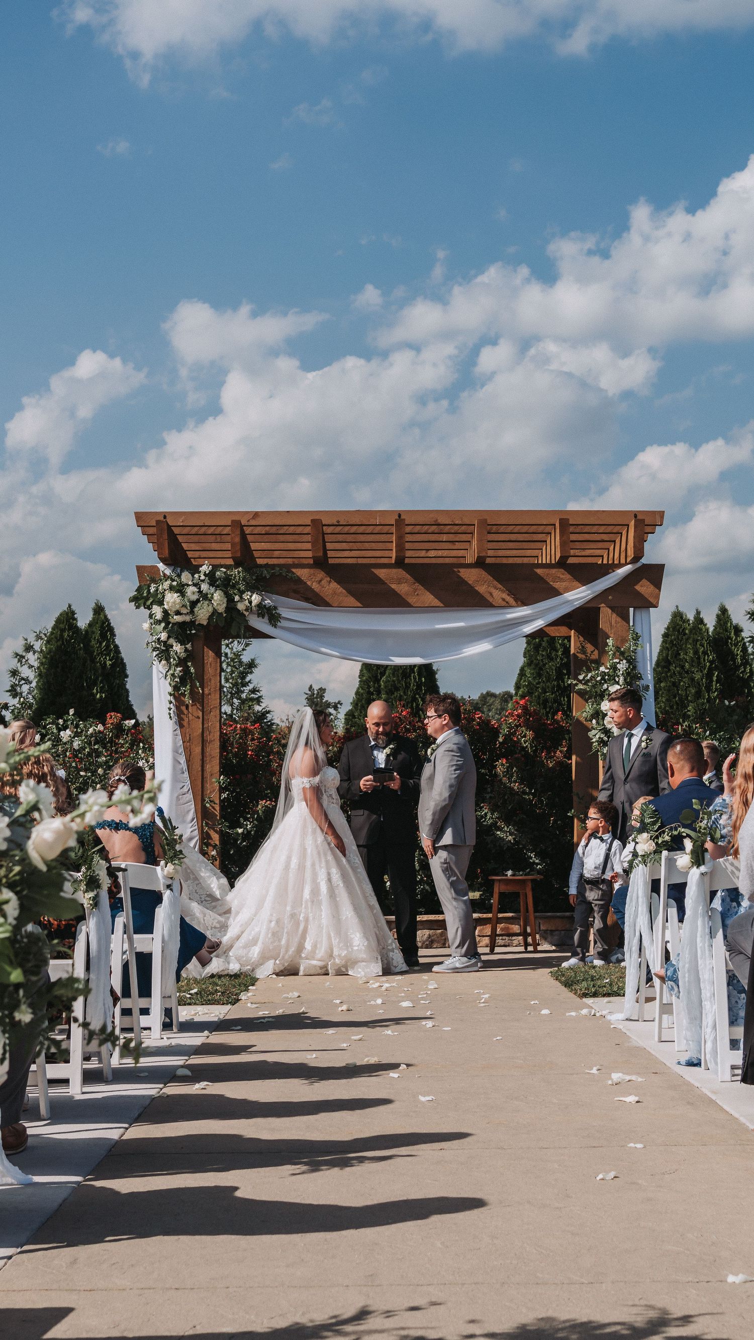 Outdoor wedding ceremony with bride in white gown at wooden pergola altar, white chairs, floral decor, and blue sky.